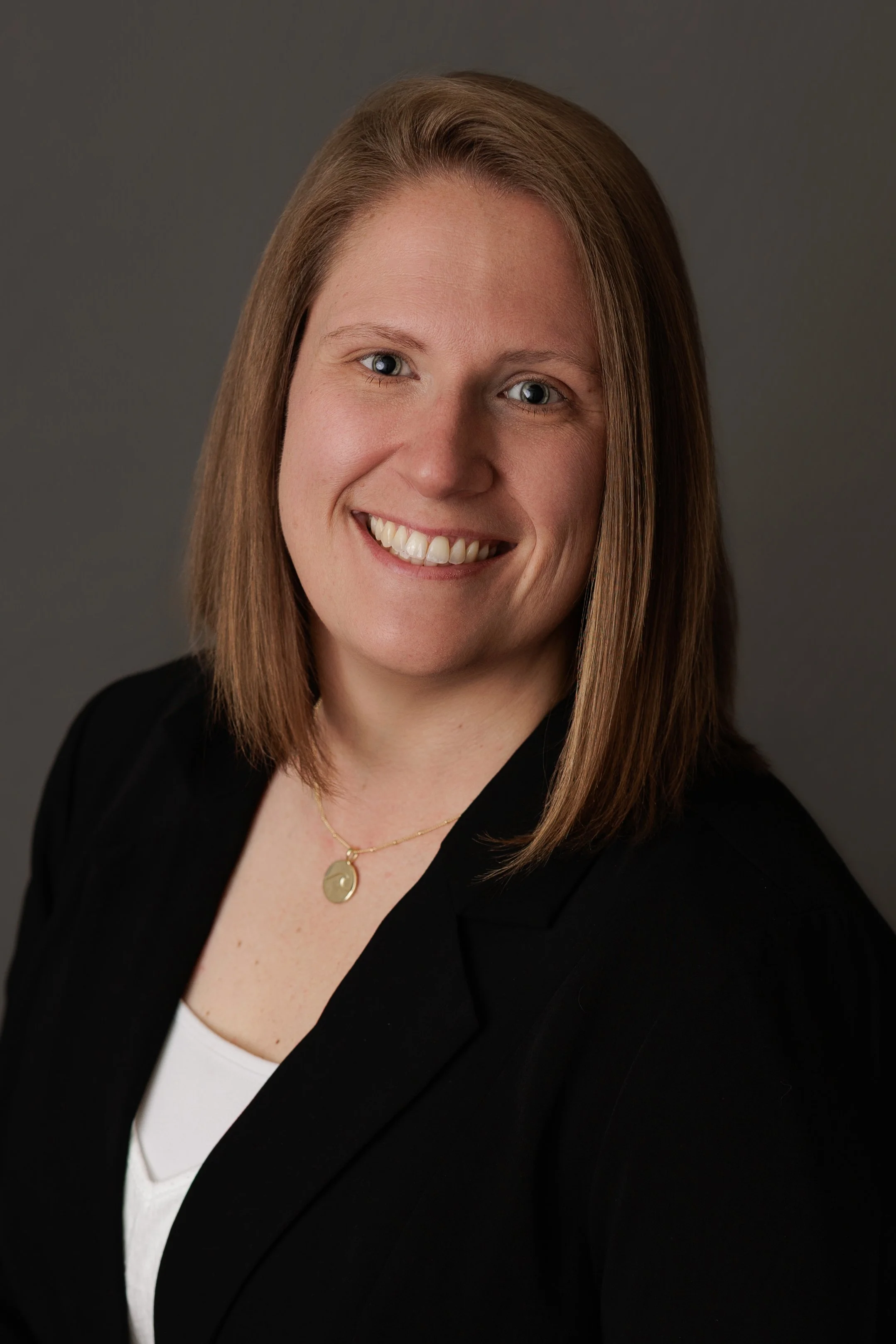 A professional woman with shoulder-length reddish-brown hair smiling, wearing a black blazer over a white top, and a gold necklace with a round pendant, against a gray background.