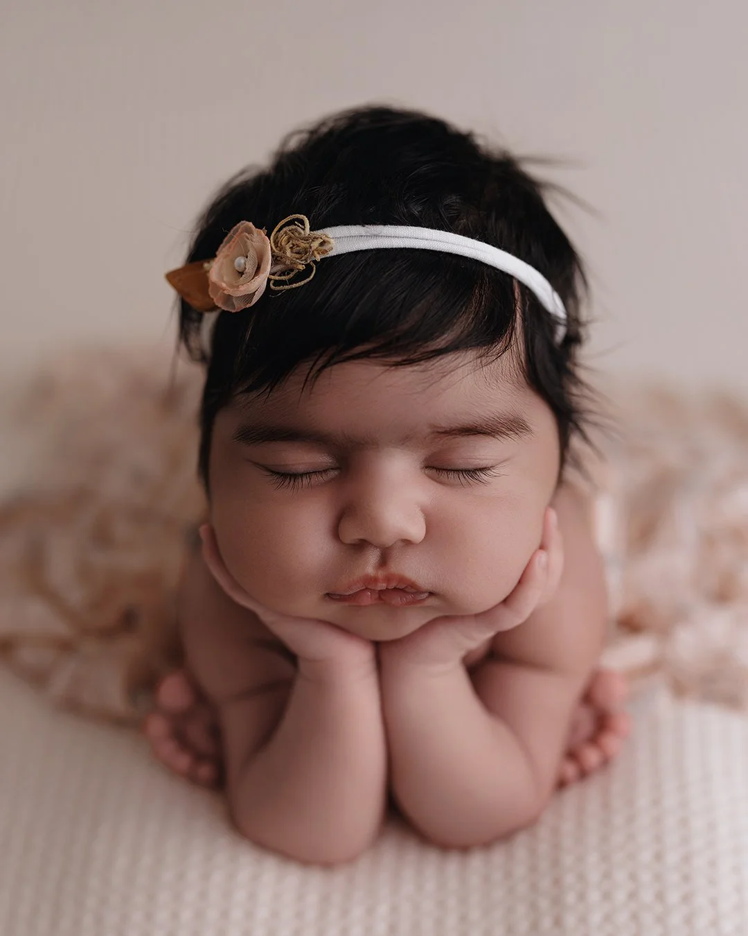 Close-up of a sleeping baby resting chin on hands, with eyes closed, wearing a headband with a flower, on a soft surface.