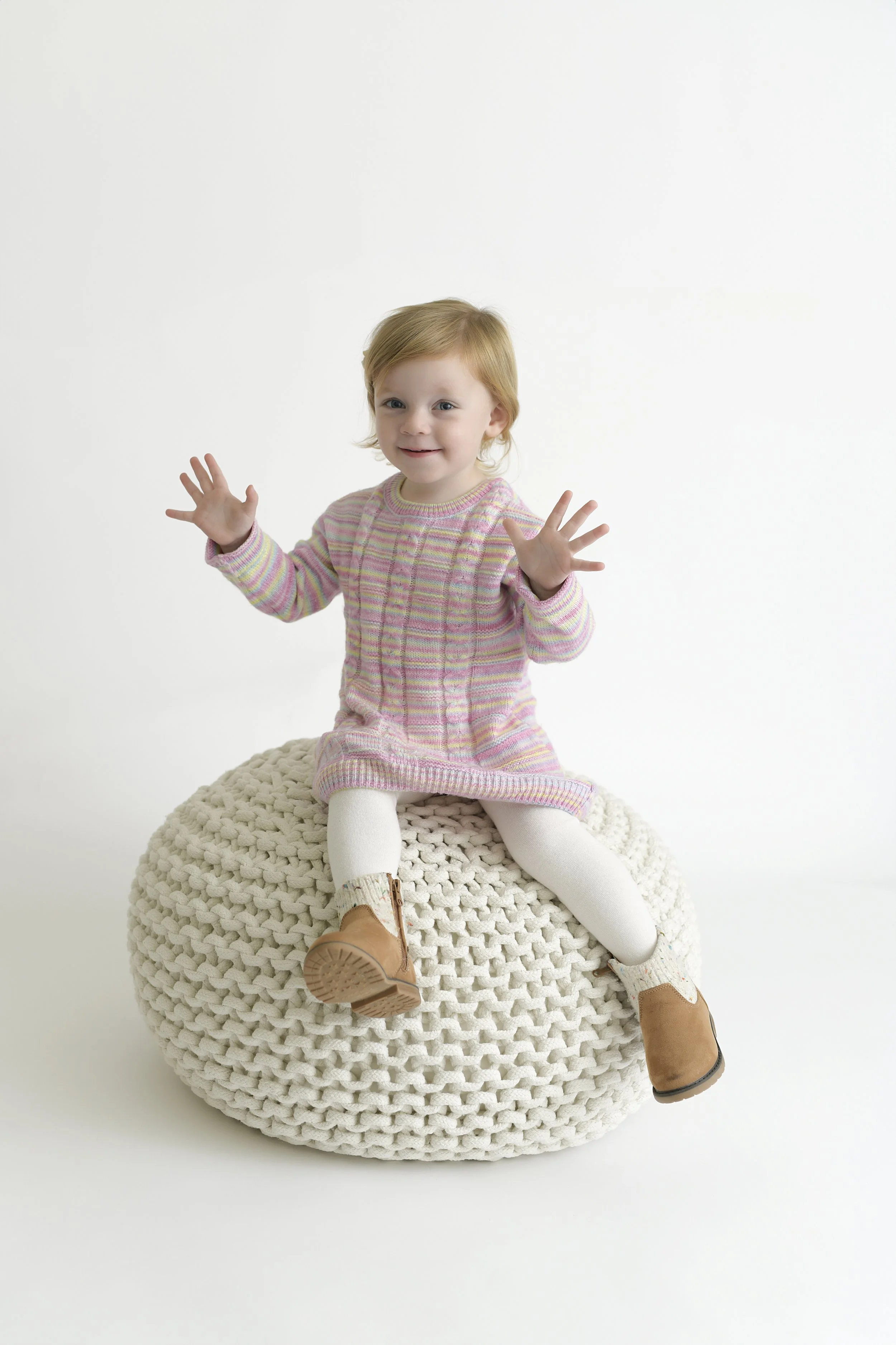 A young girl with red hair sitting on a chunky knit pouf, wearing a pastel striped sweater, white tights, and brown boots, smiling and reaching forward with her hands.