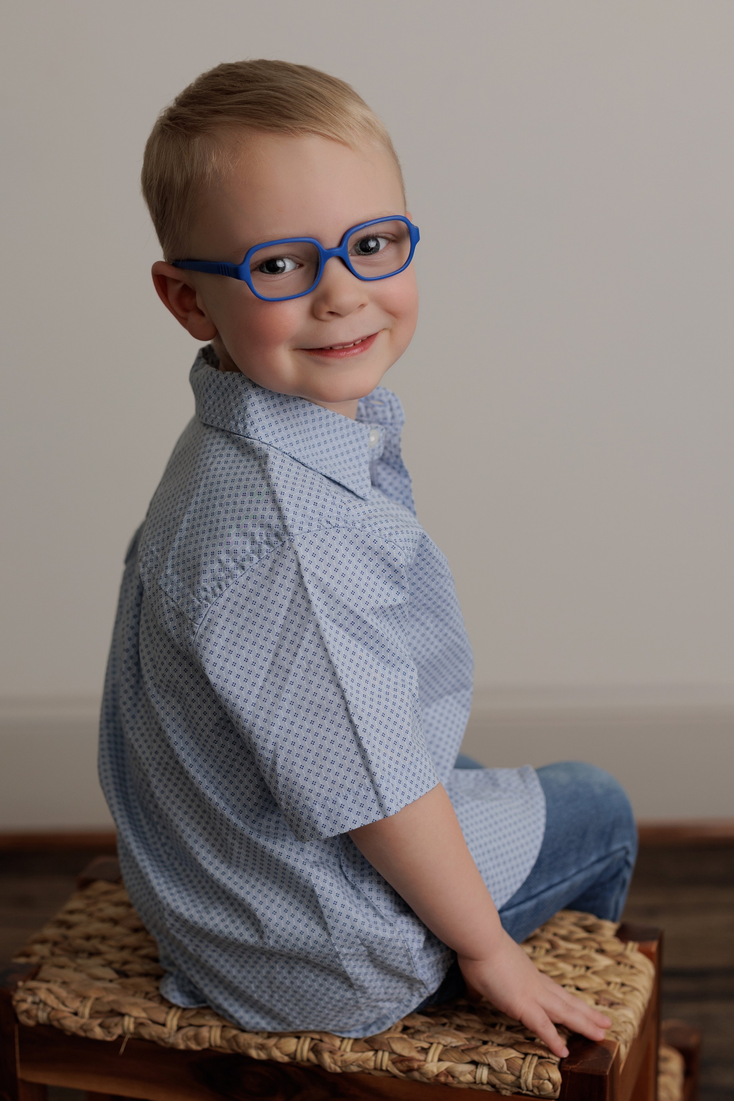A young boy sitting on a woven basket with a white wall in the background. He has blond hair, wears blue glasses, a blue polka dot shirt, and blue jeans. He is smiling and looking at the camera.