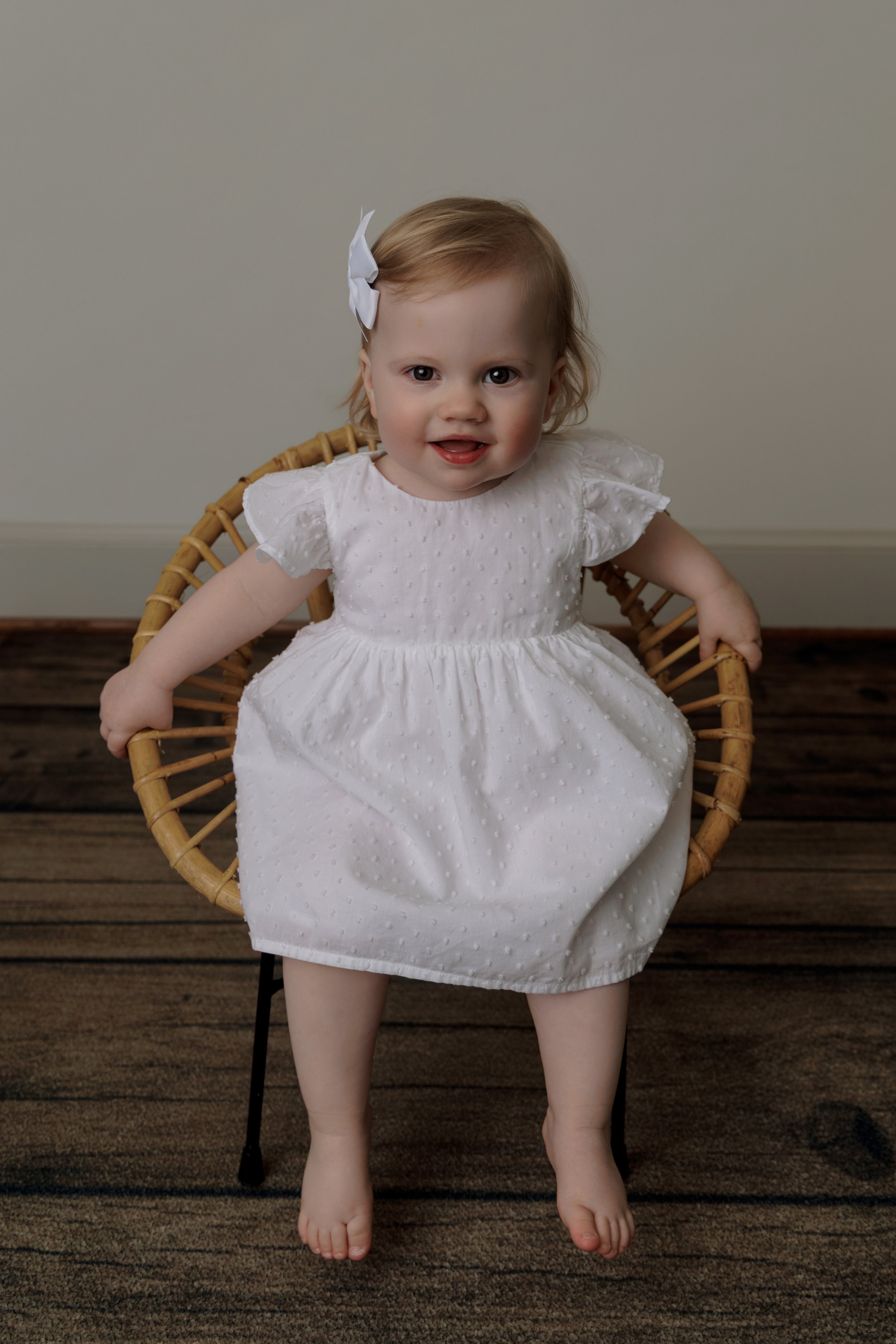 A young girl with light brown hair and a white bow, smiling while sitting in a wicker chair wearing a white dress, with her feet on a dark striped carpet and a plain wall in the background.