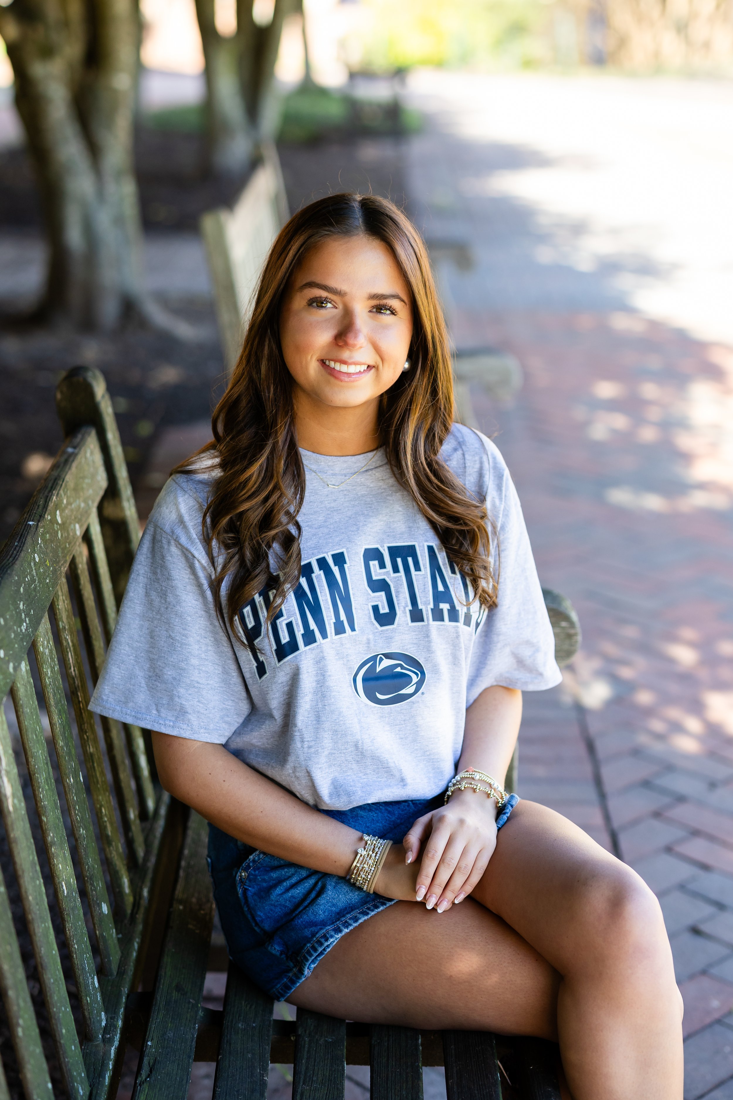 A young woman with long brown hair sitting on a wooden bench outdoors, wearing a gray Penn State sweatshirt, denim shorts, and jewelry, smiling at the camera with trees and sunlight in the background.