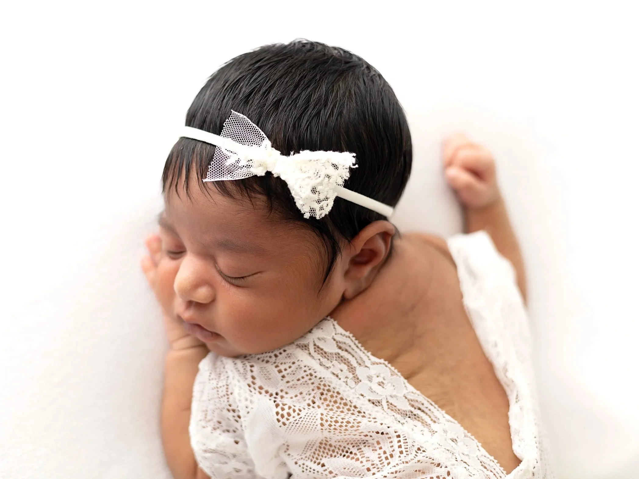 Close-up of a sleeping newborn baby with dark hair, wearing a lace white outfit and a white headband with lace and mesh details, resting on a soft white surface.