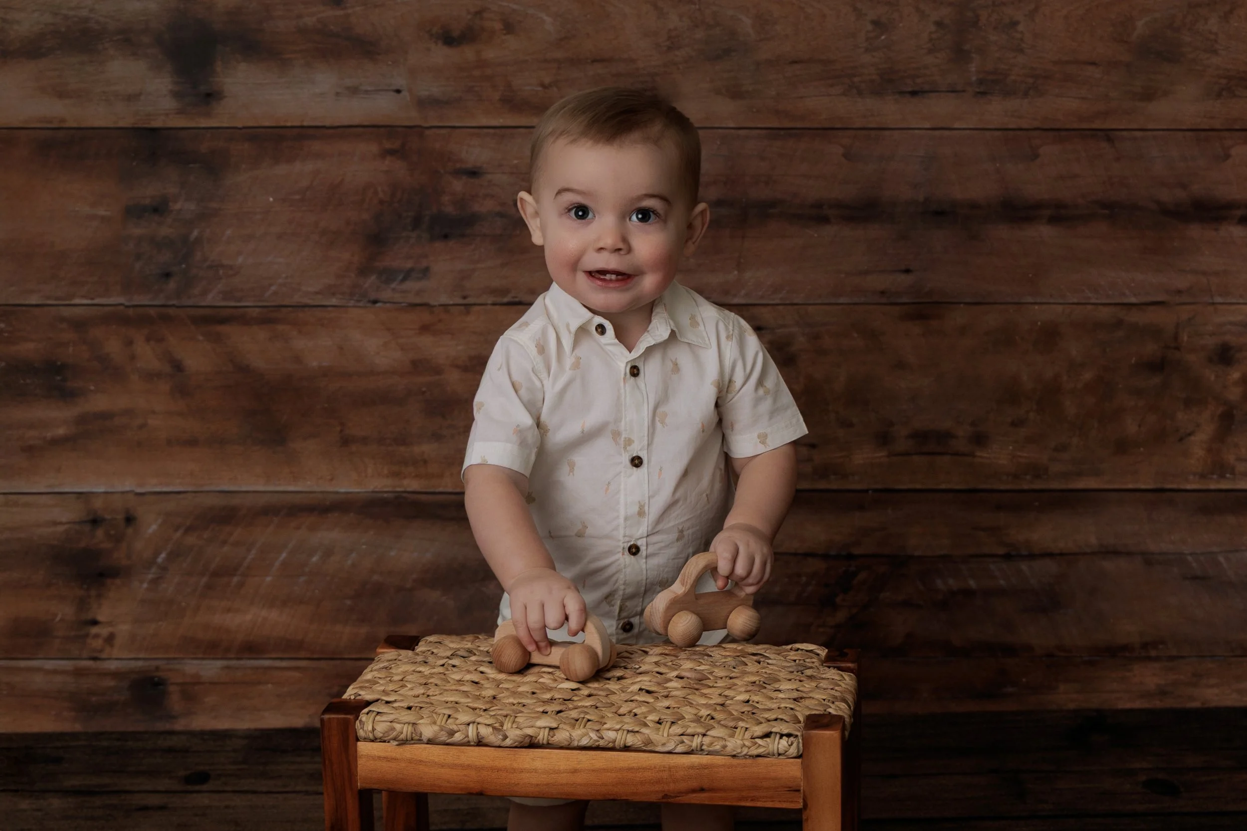 A young boy with short light brown hair, wearing a cream-colored shirt with a pattern and black buttons, sitting on a woven stool against a wooden wall background, holding a wooden toy roller.