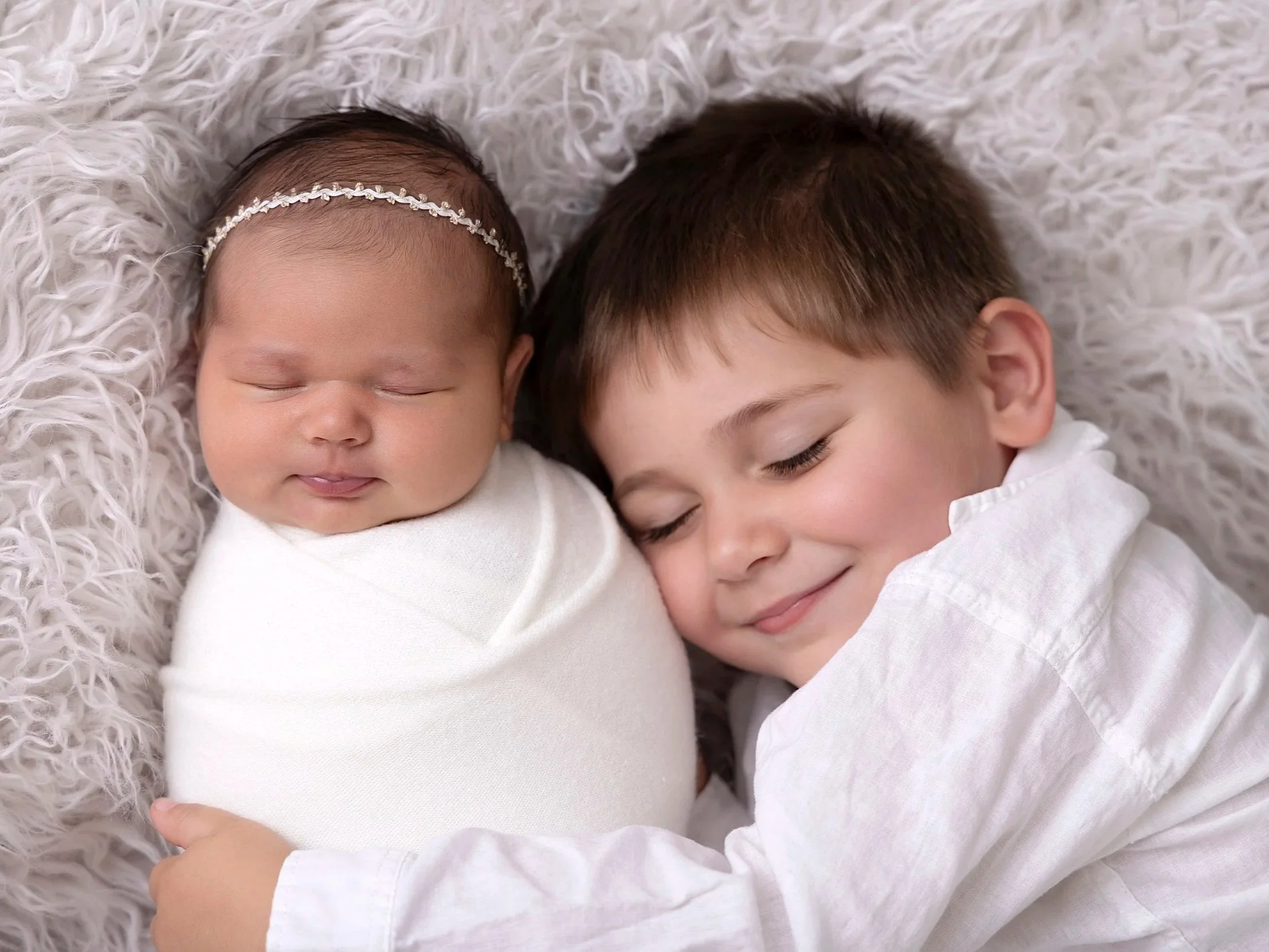 A young boy and a newborn baby girl lying close together on a fluffy white blanket, both with their eyes closed and smiling softly.