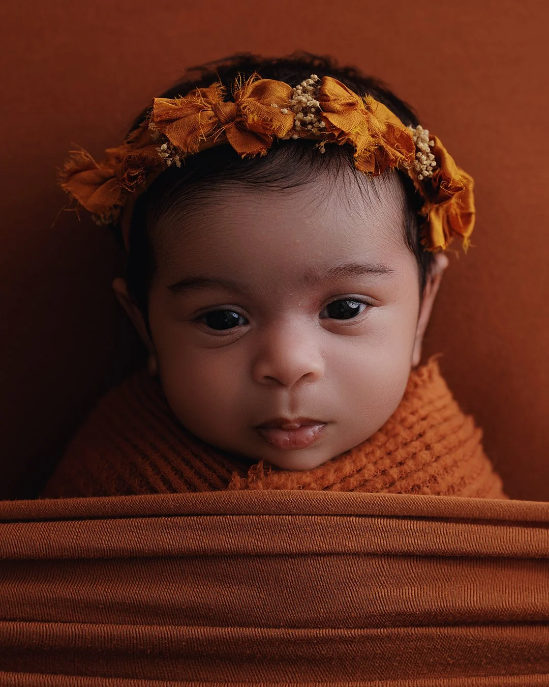 Close-up portrait of a baby wearing a brown knitted outfit and a yellow floral headband, lying on a brown surface.