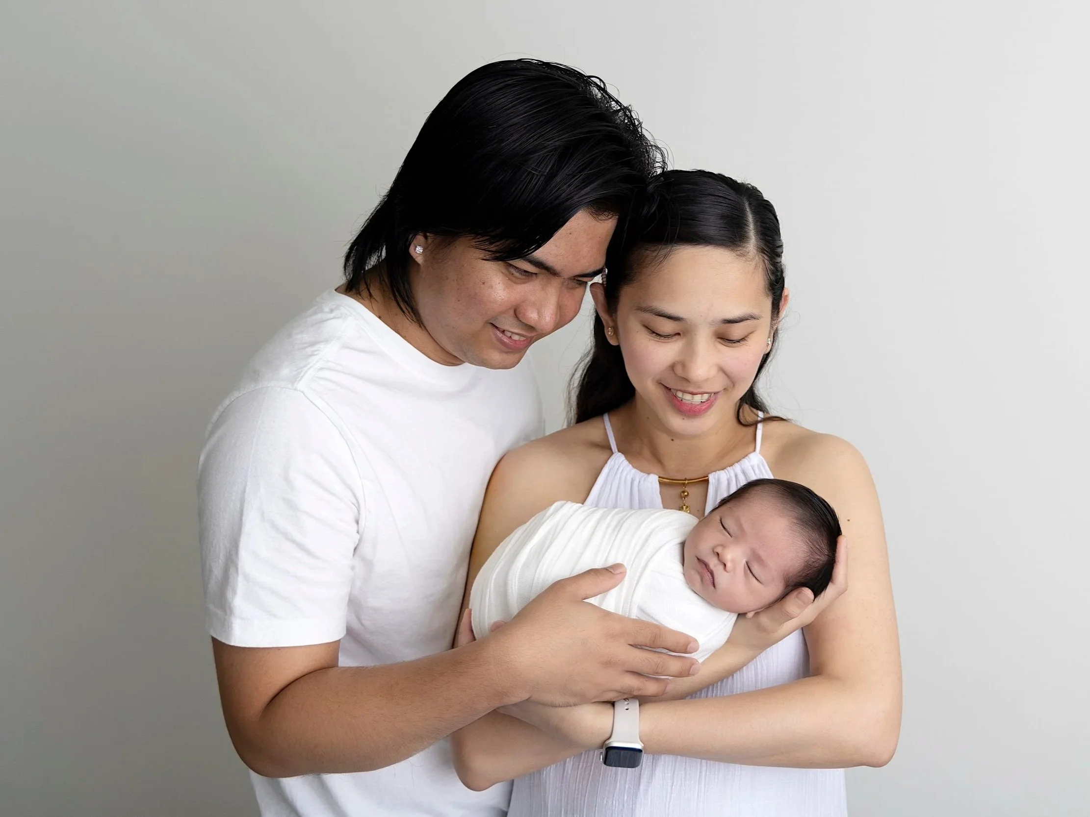 A happy family with a man and woman holding a newborn baby, all smiling, standing against a plain white background.