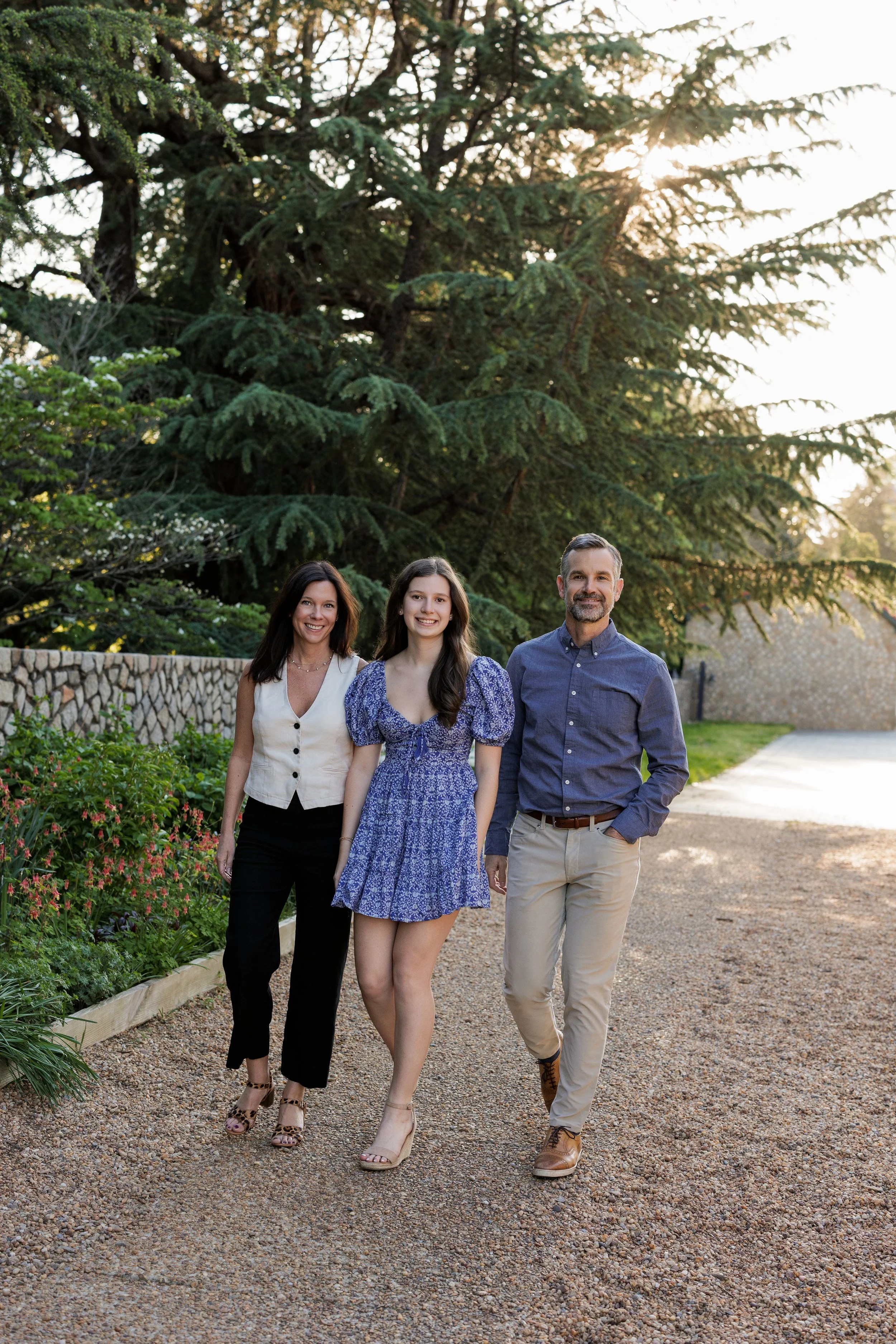 A family of three walking outdoors on a gravel path, with large trees and a stone wall in the background during late afternoon or early evening.