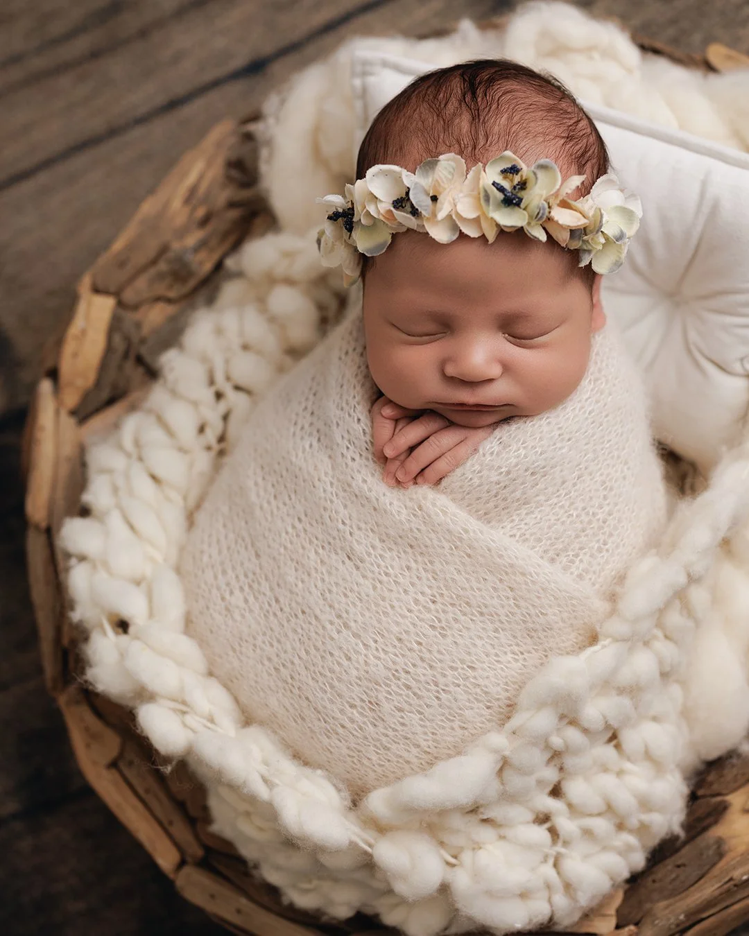 A sleeping newborn baby wrapped in a cream-colored knitted blanket, lying in a round wooden basket lined with soft cotton and wool, wearing a floral headband with white petals and small dark accents.