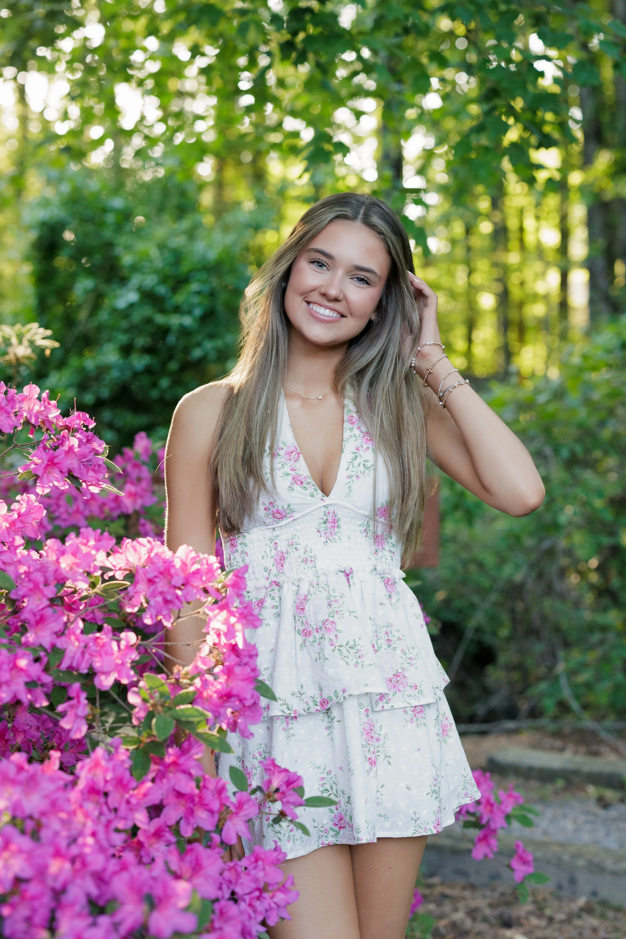 A young woman with long, light brown hair smiling outdoors near pink flowering bushes and green trees.
