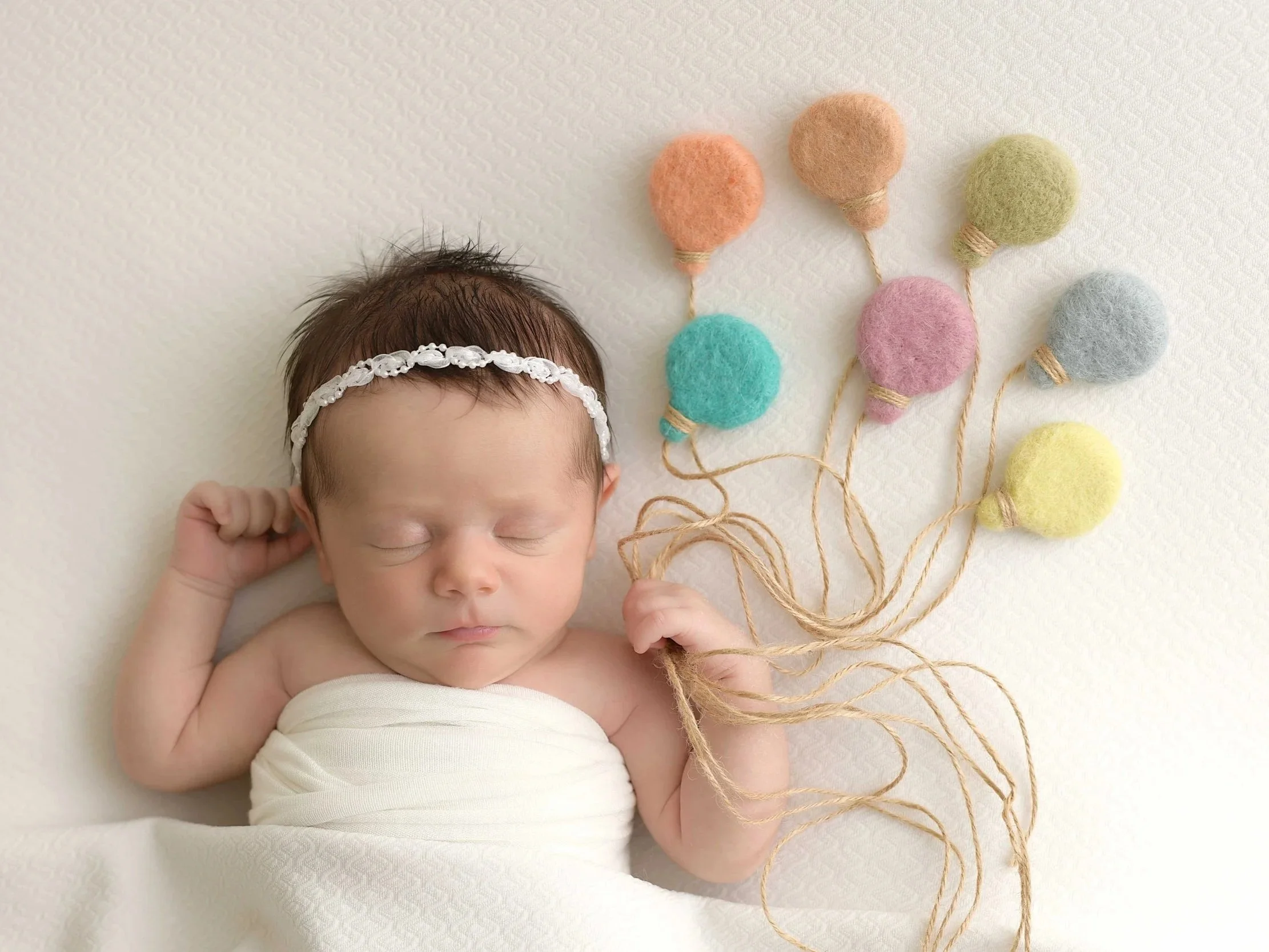 A sleeping newborn baby lying on a white textured surface, wearing a white headband, holding felted balloon strings with colorful felted balloons attached.