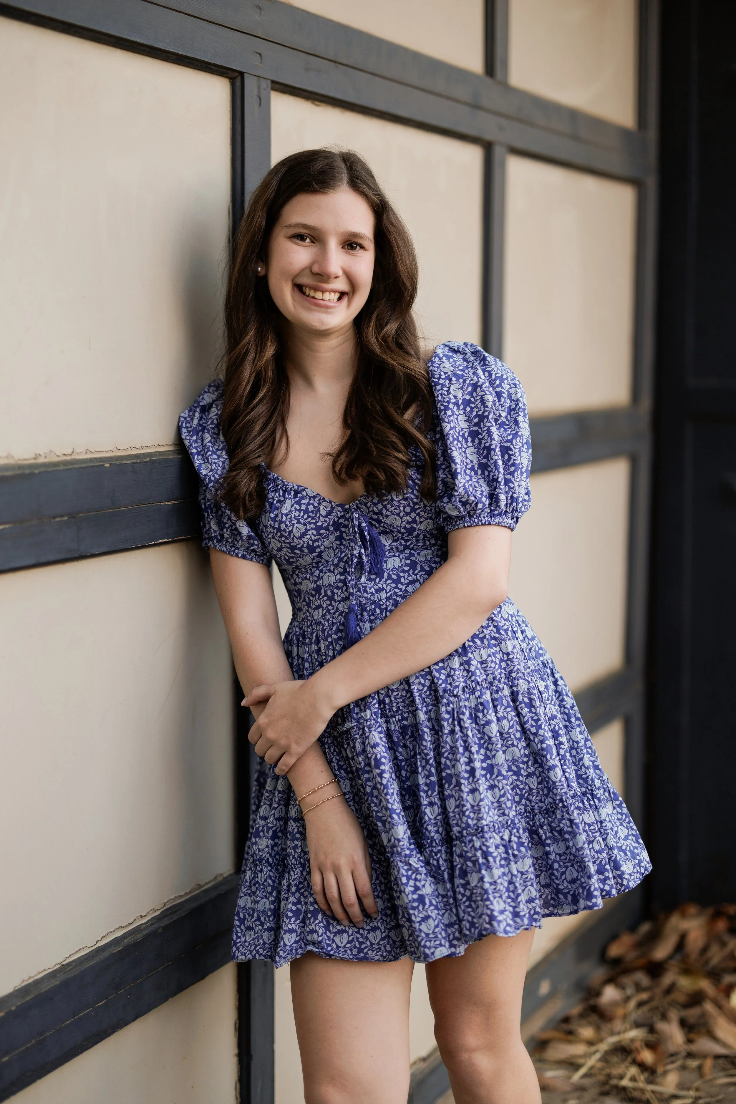 A young woman in a blue floral dress standing against a beige and black wall, smiling at the camera.