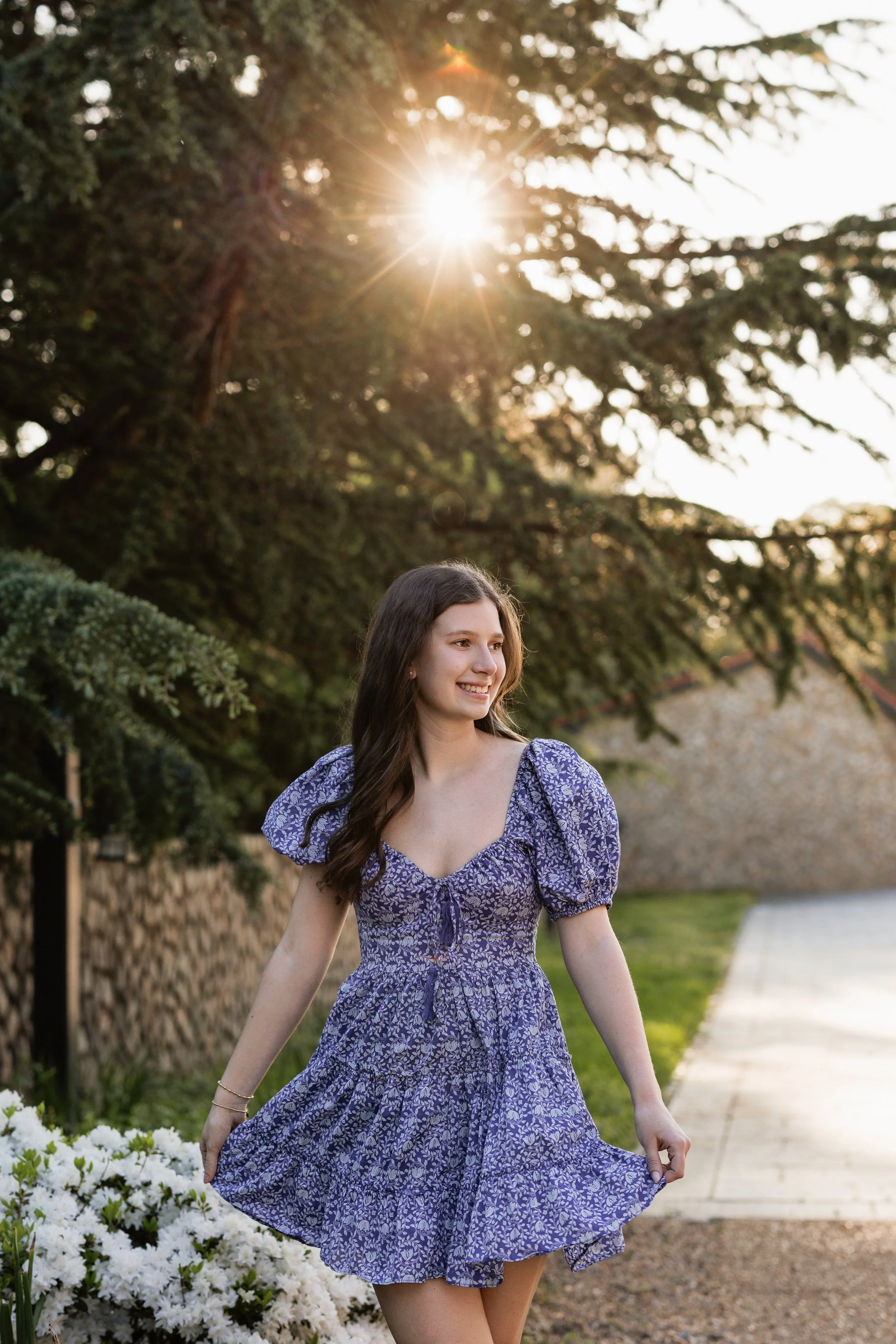 A young woman in a purple floral dress walking outdoors during sunset, with sunlight filtering through tree branches in the background.