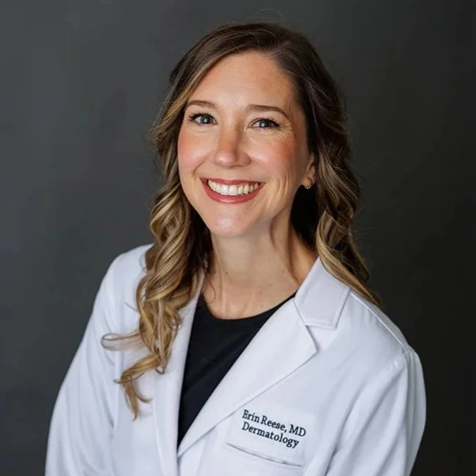A smiling woman in a white medical coat with her name and title, standing against a gray background.