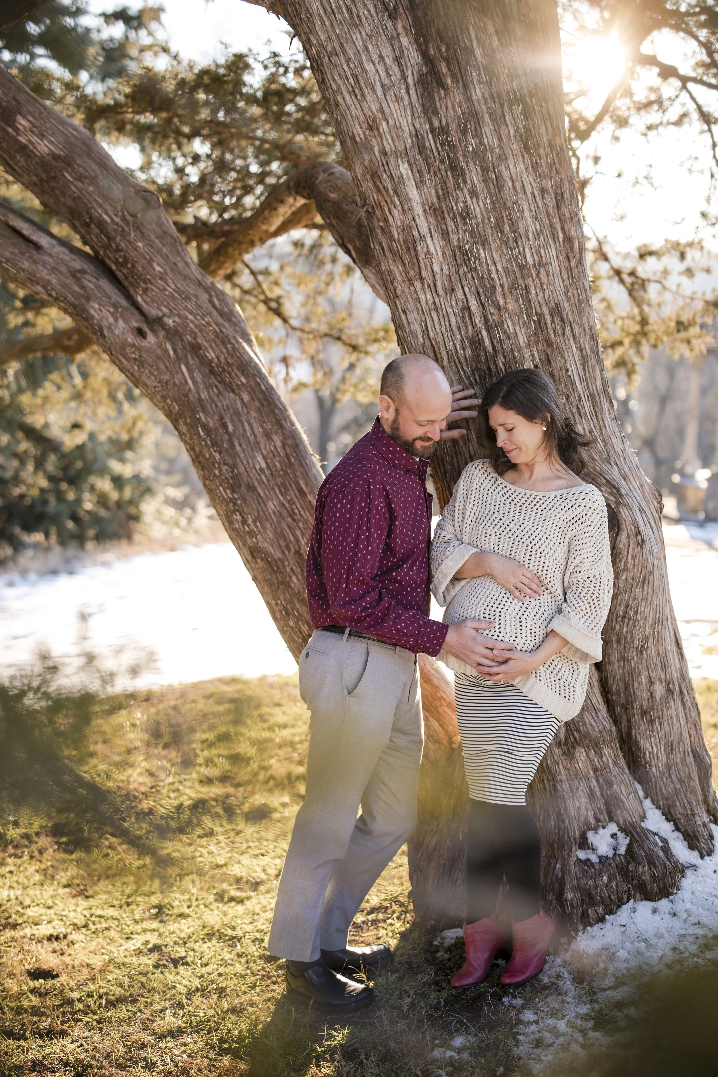 A couple, a man and a pregnant woman, standing outdoors near a large tree with bare branches, embracing and touching their bellies as they look at each other. The sun is setting behind them, casting a warm glow, with snow on the ground.