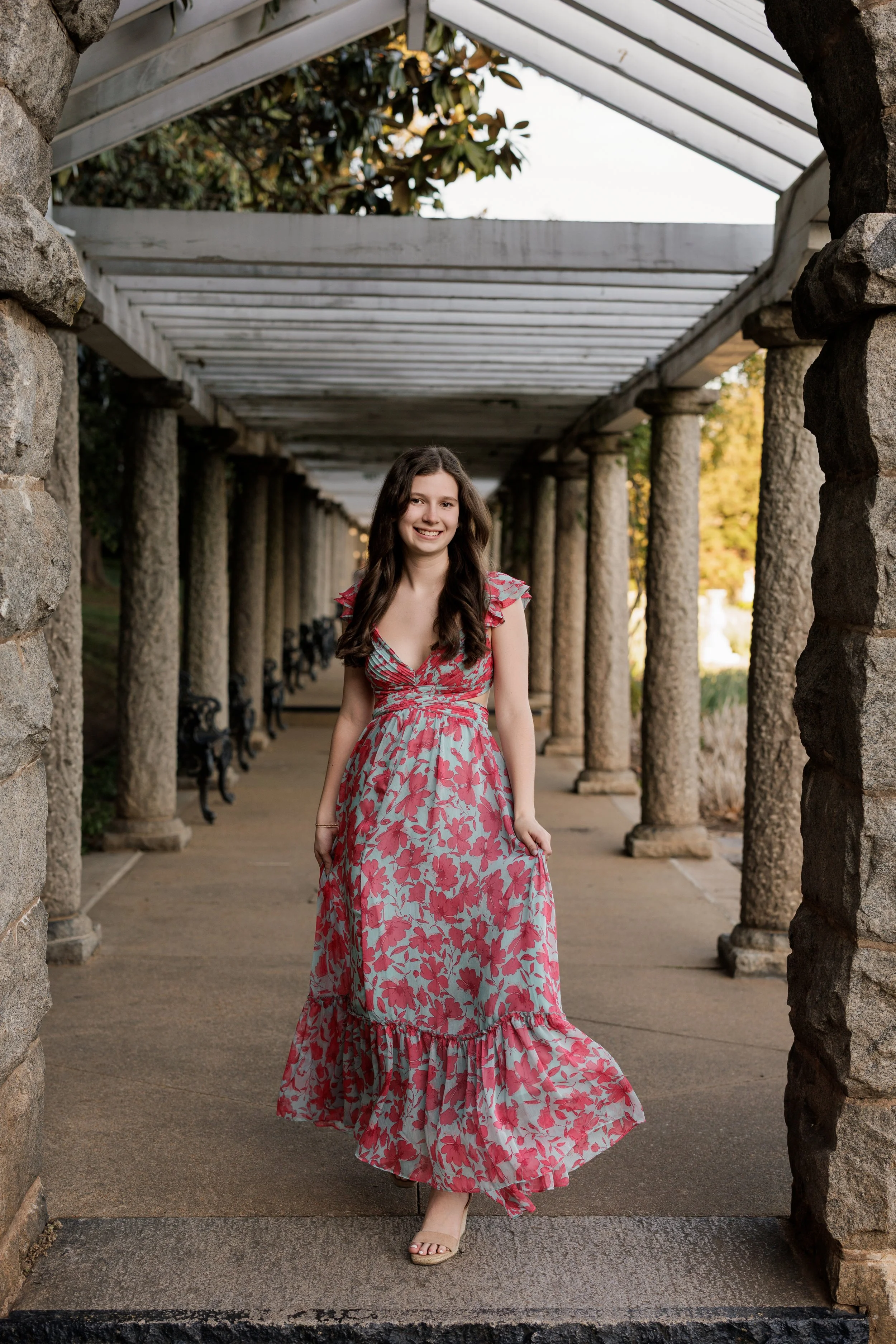 A young woman wearing a long, pink floral dress, standing under a stone and wood pergola with columns, in an outdoor setting during daylight. Trees with autumn leaves are visible in the background.