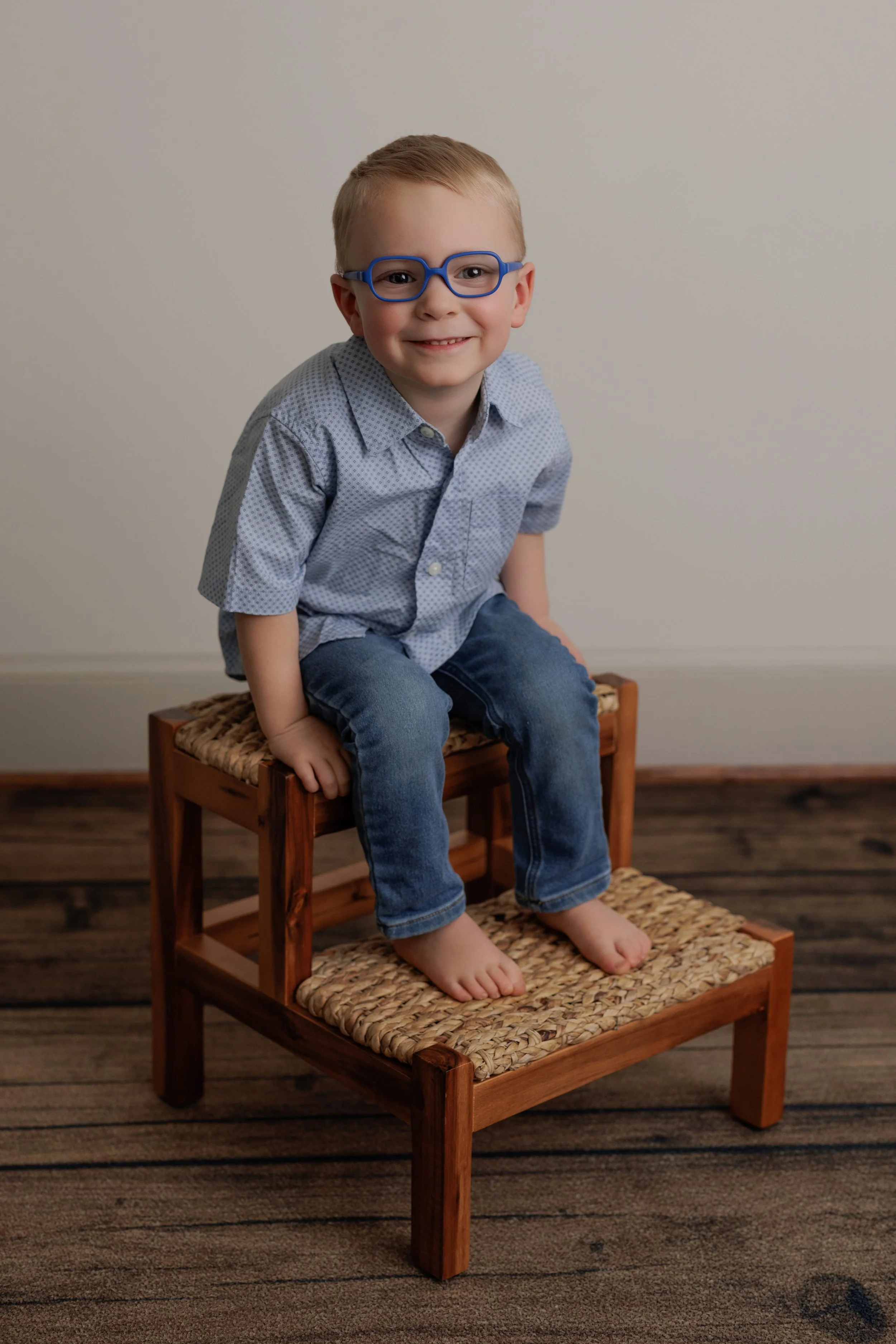 A young boy with blonde hair, wearing blue glasses, a light blue button-up shirt, and jeans, sitting barefoot on a woven rattan stool with another matching stool beneath his feet, against a plain beige wall with wooden floor.