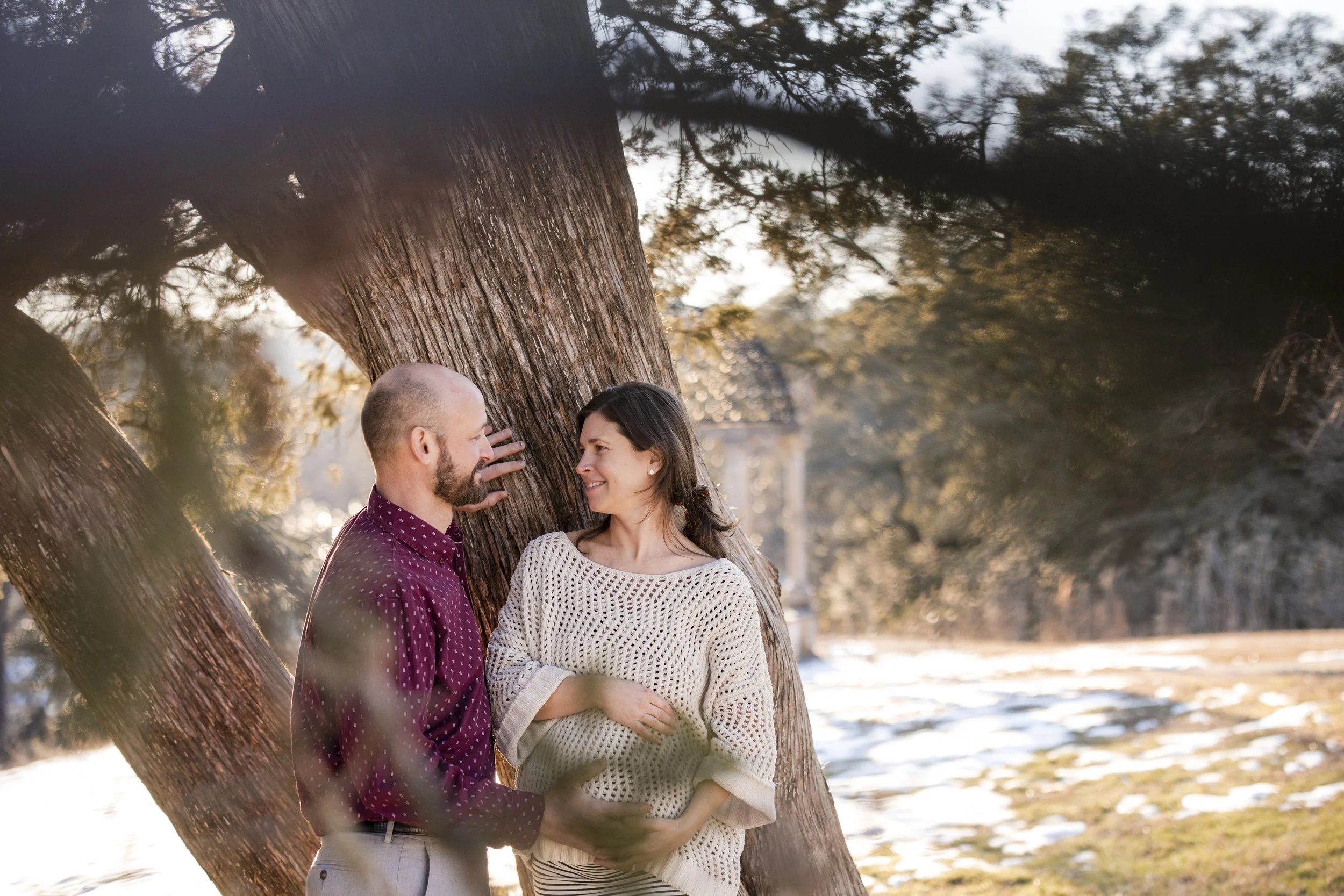 A man and woman standing close together near a large tree outdoors, smiling and looking at each other in a park setting during late afternoon or early evening.