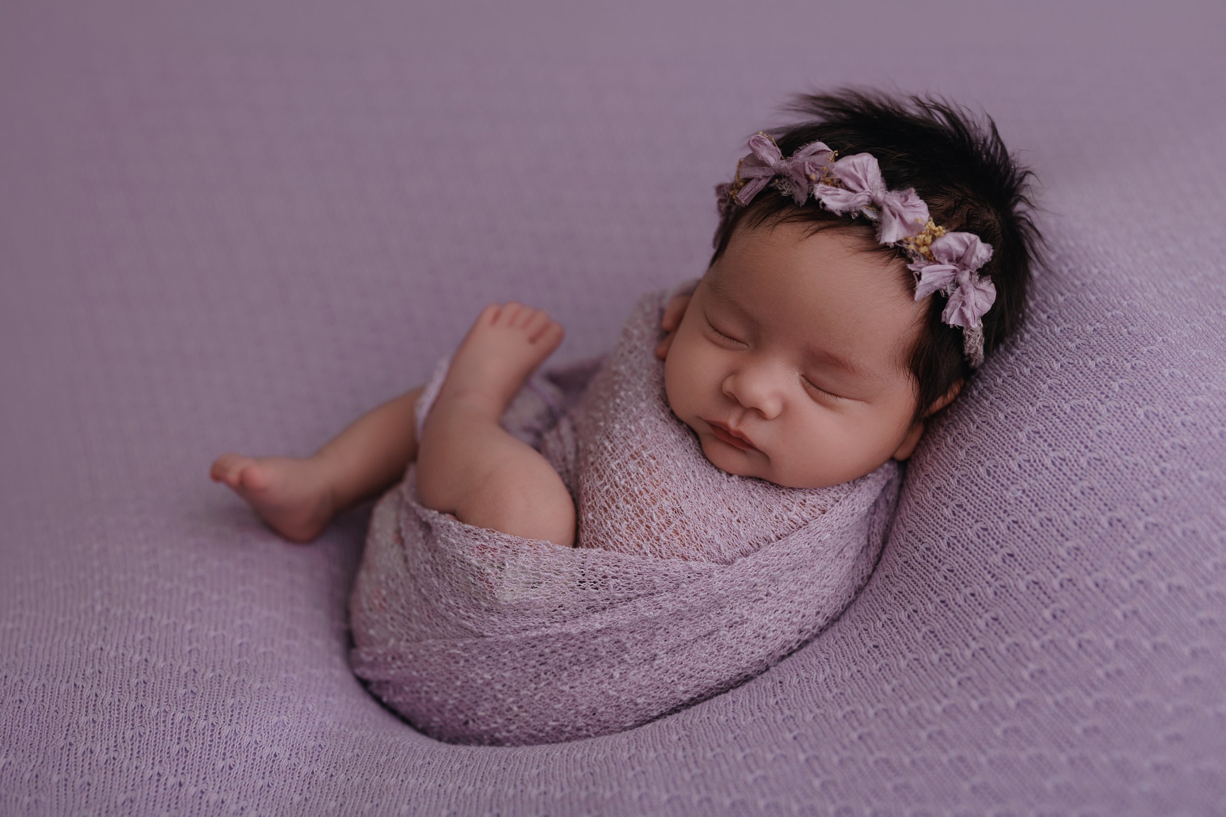 A sleeping infant wrapped in a pink knitted blanket, lying on a soft pink surface, wearing a floral headband with pink and purple fabric flowers.