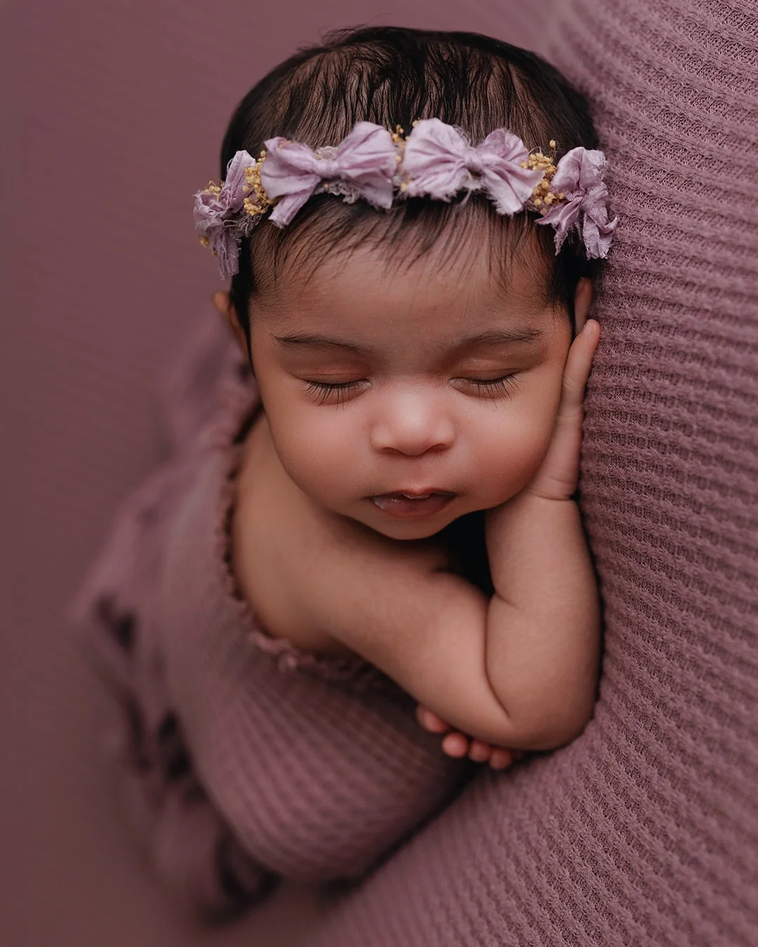 A sleeping baby girl with a purple flower headband, resting her head on a pink textured surface, with her eyes closed and a peaceful expression.