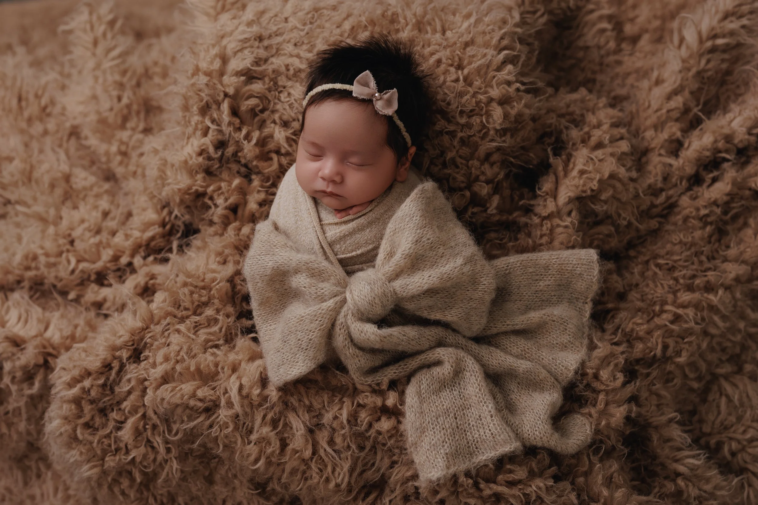 A sleeping baby girl with a beige bow headband and wrapped in a beige blanket, lying on a fluffy brown rug.