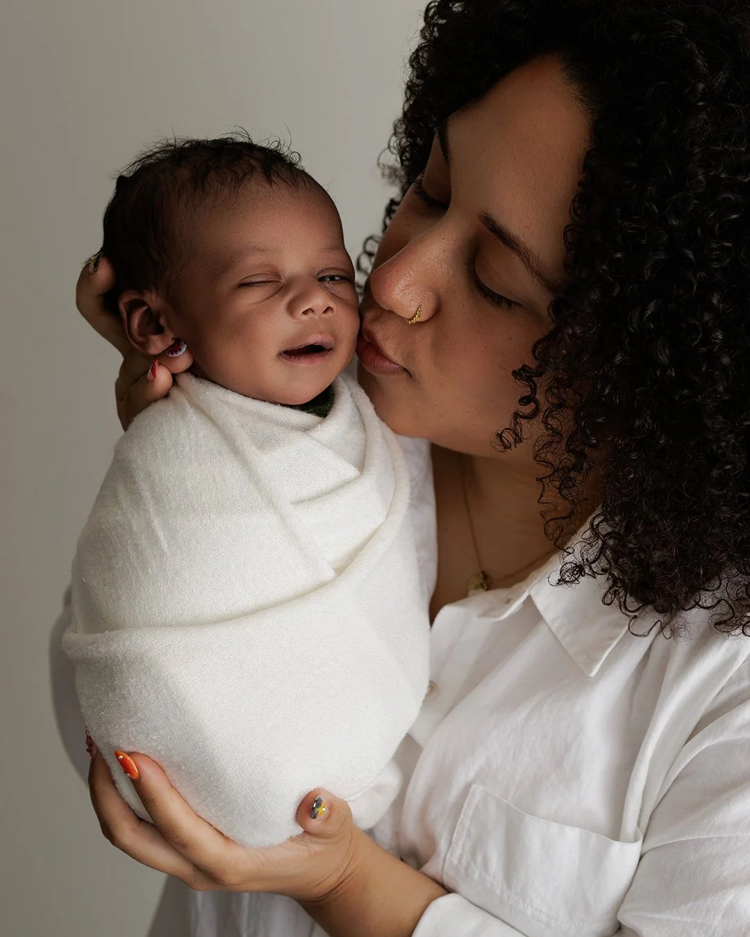 A woman with curly hair and a nose ring holds a baby wrapped in a white blanket and kisses the baby's cheek.