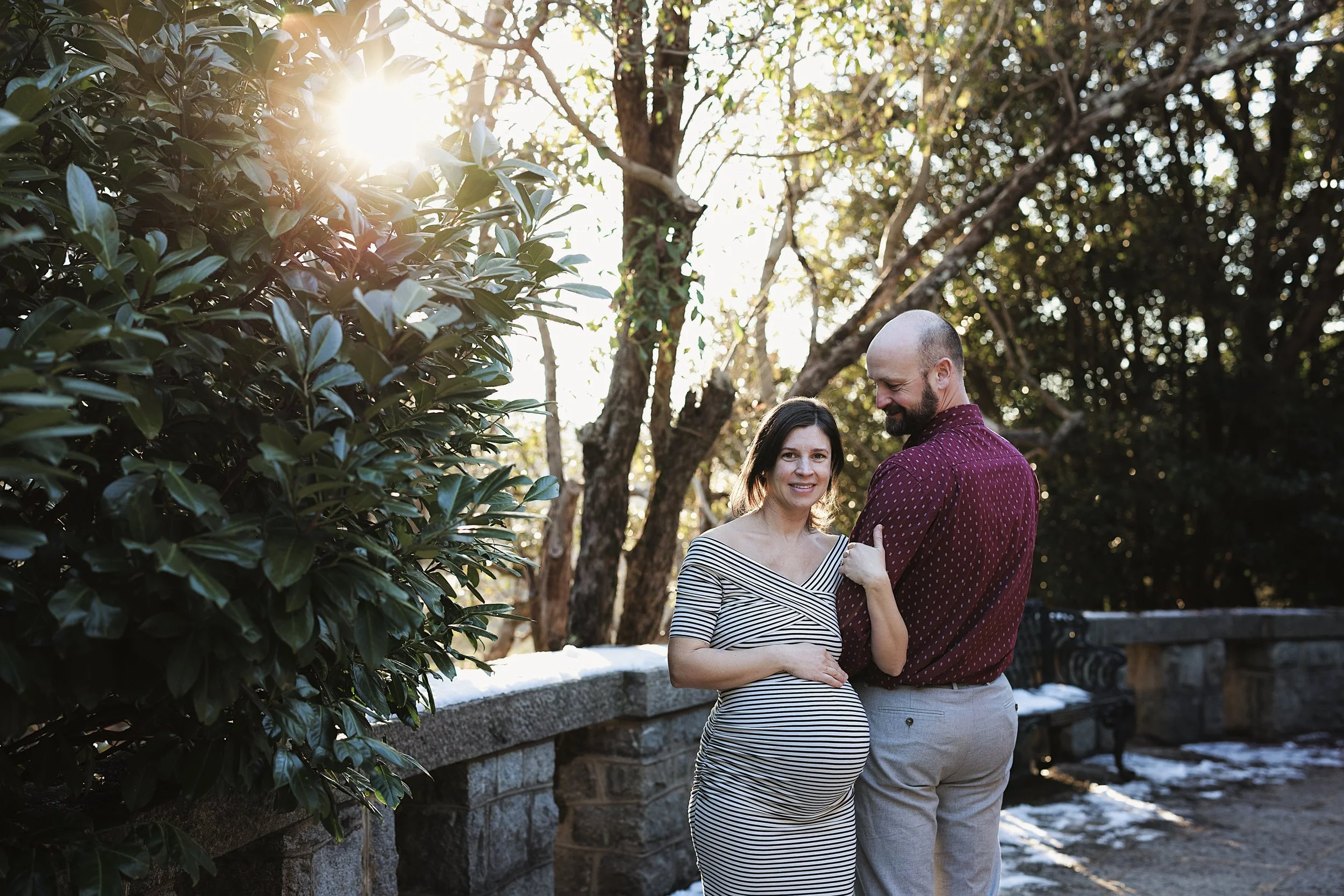 A pregnant woman and a man are standing close together outdoors near a stone railing, smiling. The woman is wearing a black and white striped dress, and the man is wearing a maroon shirt with small patterns and grey pants. Sunlight filters through tr