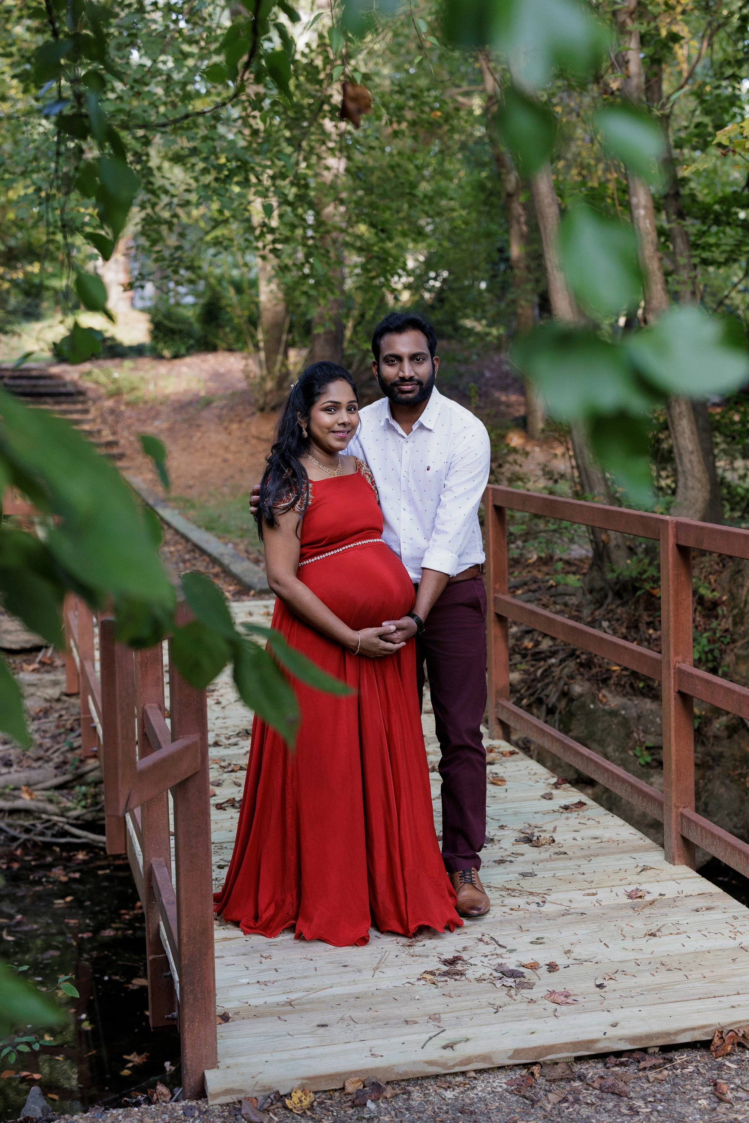 A pregnant woman in a red gown and a man in a white shirt and maroon pants standing on a wooden bridge surrounded by trees.