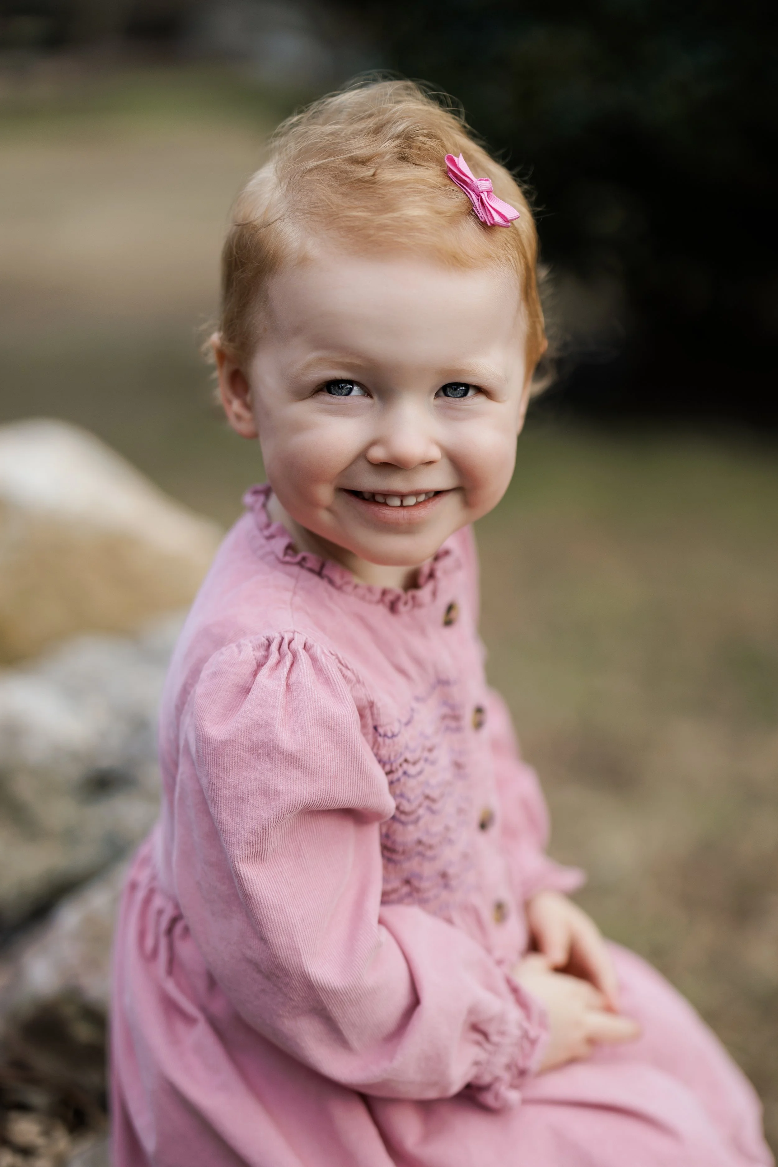 A young girl with red hair, a pink bow clip, and blue eyes, smiling while sitting outdoors on a rock or stone surface near water, wearing a pink dress with embroidered details.