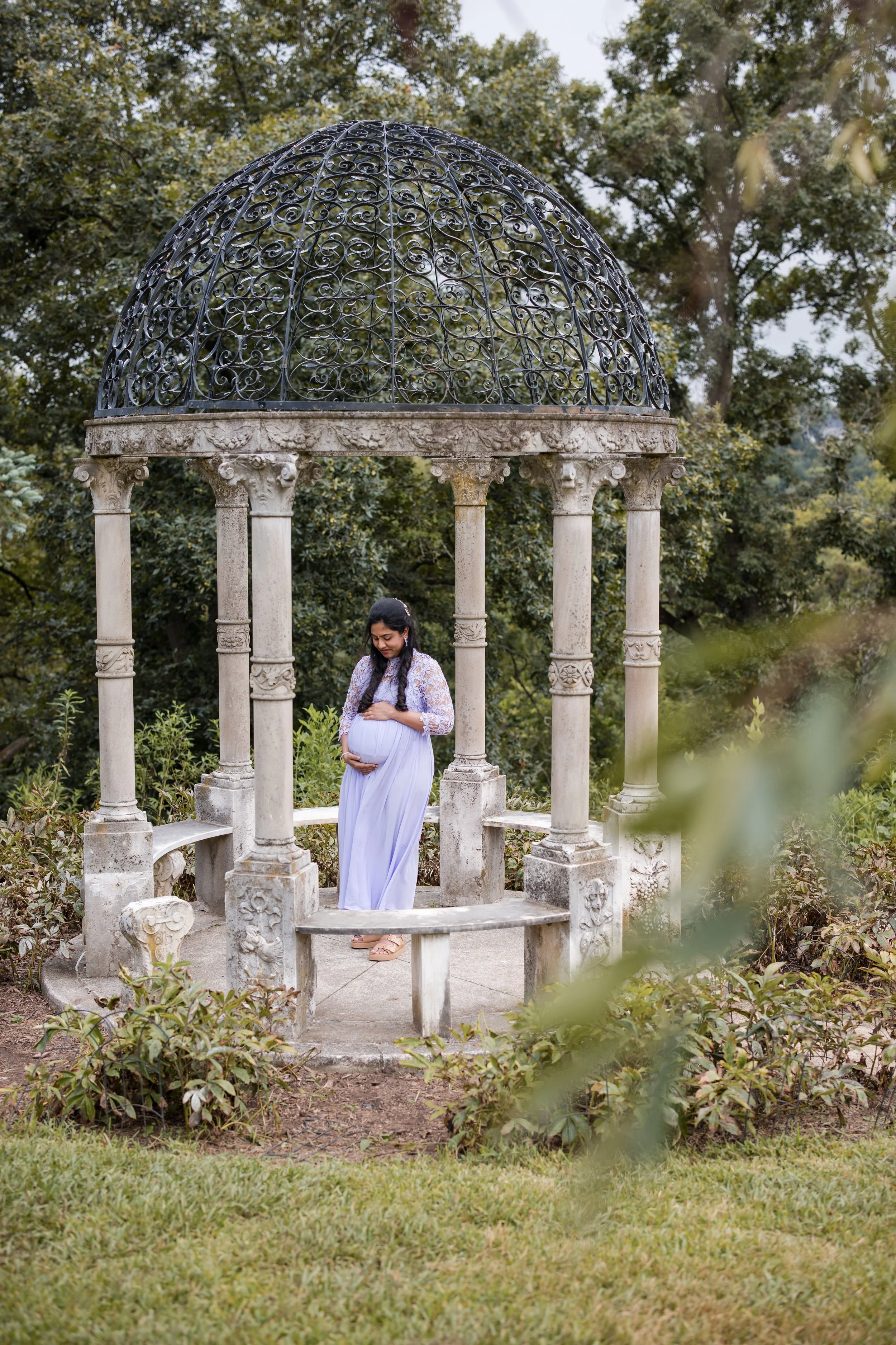 A pregnant woman standing inside a decorative stone gazebo with a metal domed roof, surrounded by lush greenery, looking down at her baby bump.