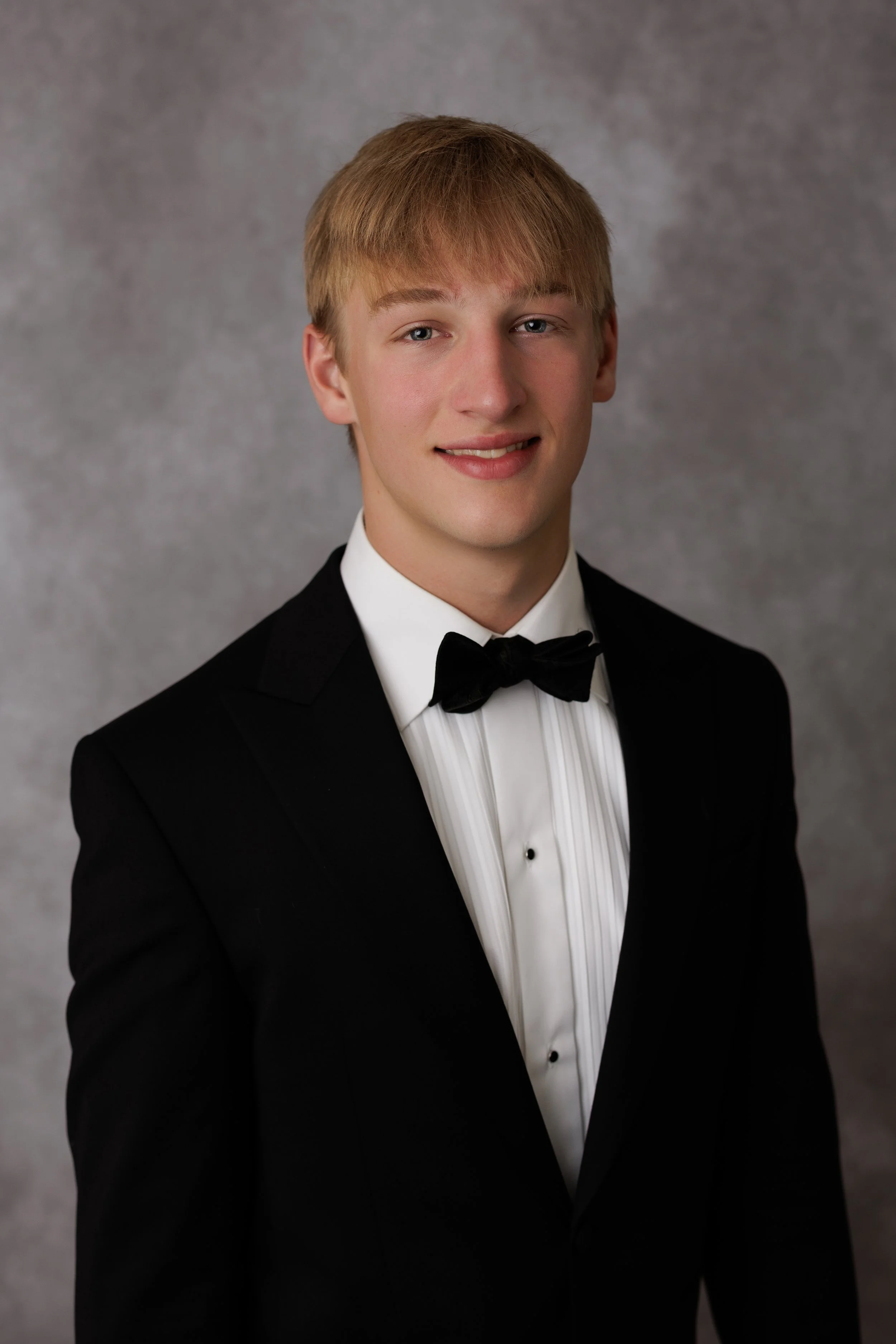 Young man with blond hair wearing a black tuxedo and black bow tie, smiling slightly, against a gray textured background.