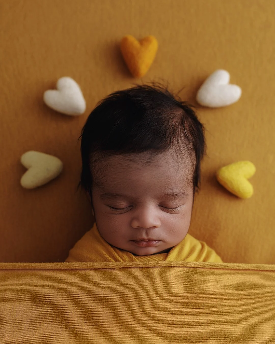 A sleeping baby with dark hair lying on a yellow blanket surrounded by small heart-shaped pillows in yellow and white colors.