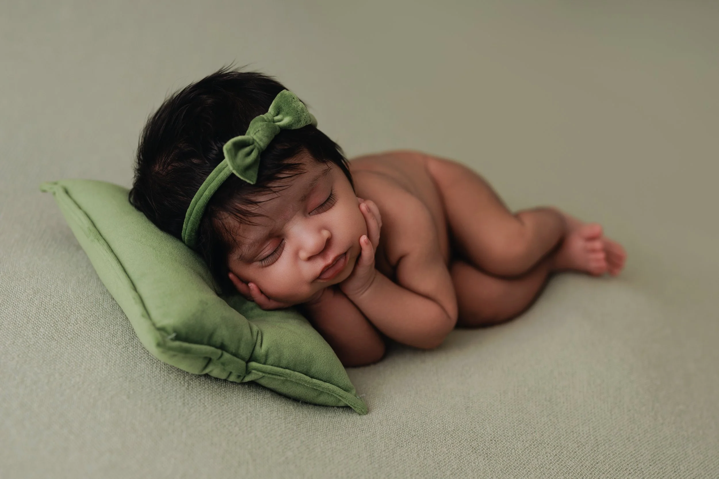 A sleeping baby with dark hair wearing a green headband with a bow, lying on a green pillow on a light-colored surface.
