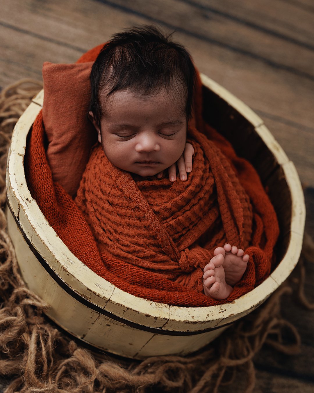 A sleeping baby wrapped in orange cloth, lying in a yellow wooden bowl with a pillow behind the head, on a rustic wooden floor.
