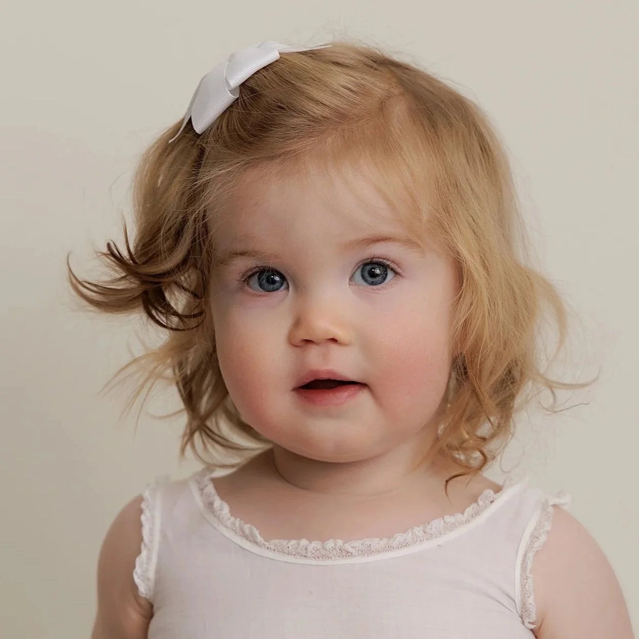 Close-up of a young girl with red hair, blue eyes, wearing a white sleeveless top with lace trim and a white bow in her hair, against a plain background.