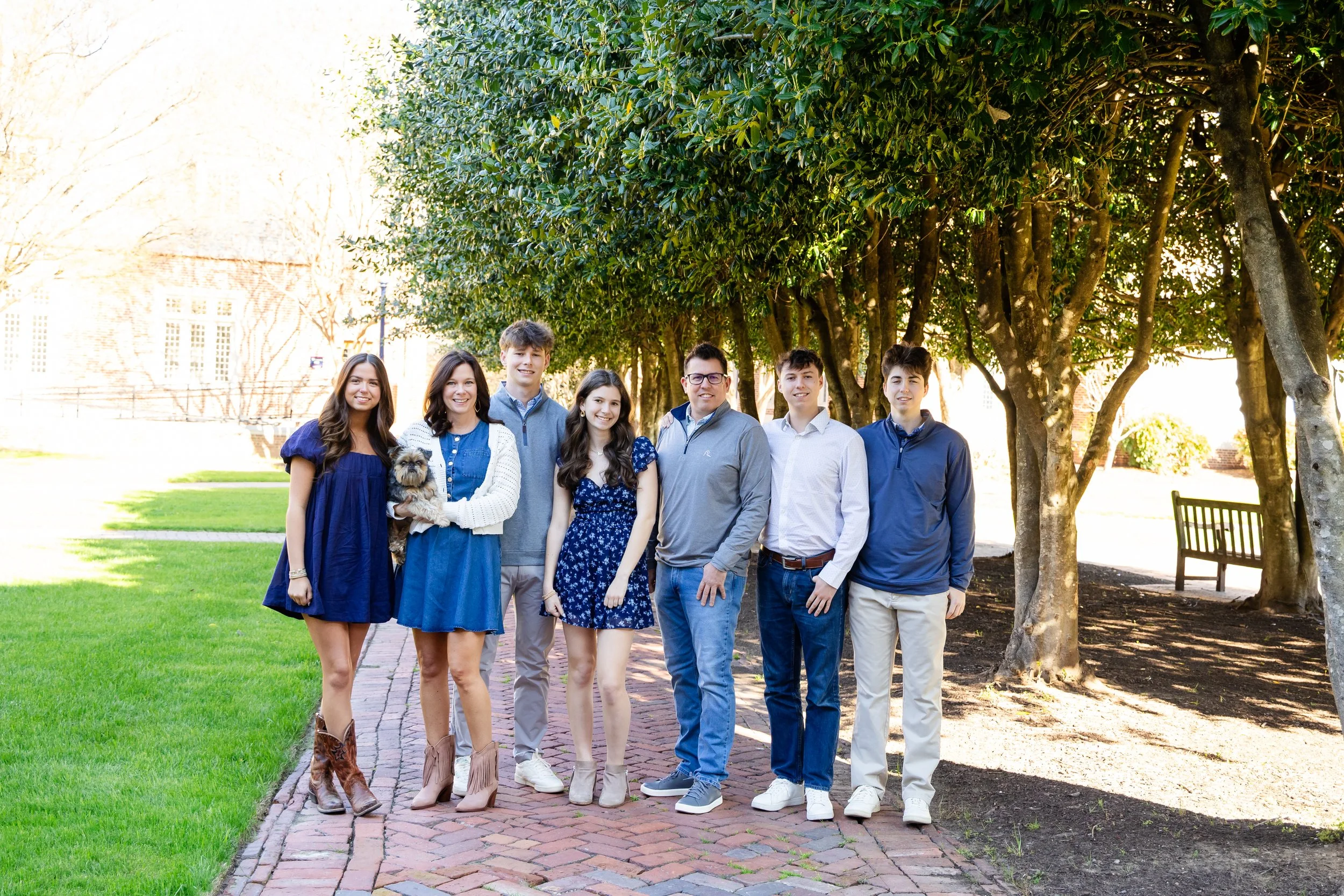 A group of nine teenagers standing outdoors on a brick sidewalk, smiling, with trees and a grassy area in the background. One girl is holding a small dog.