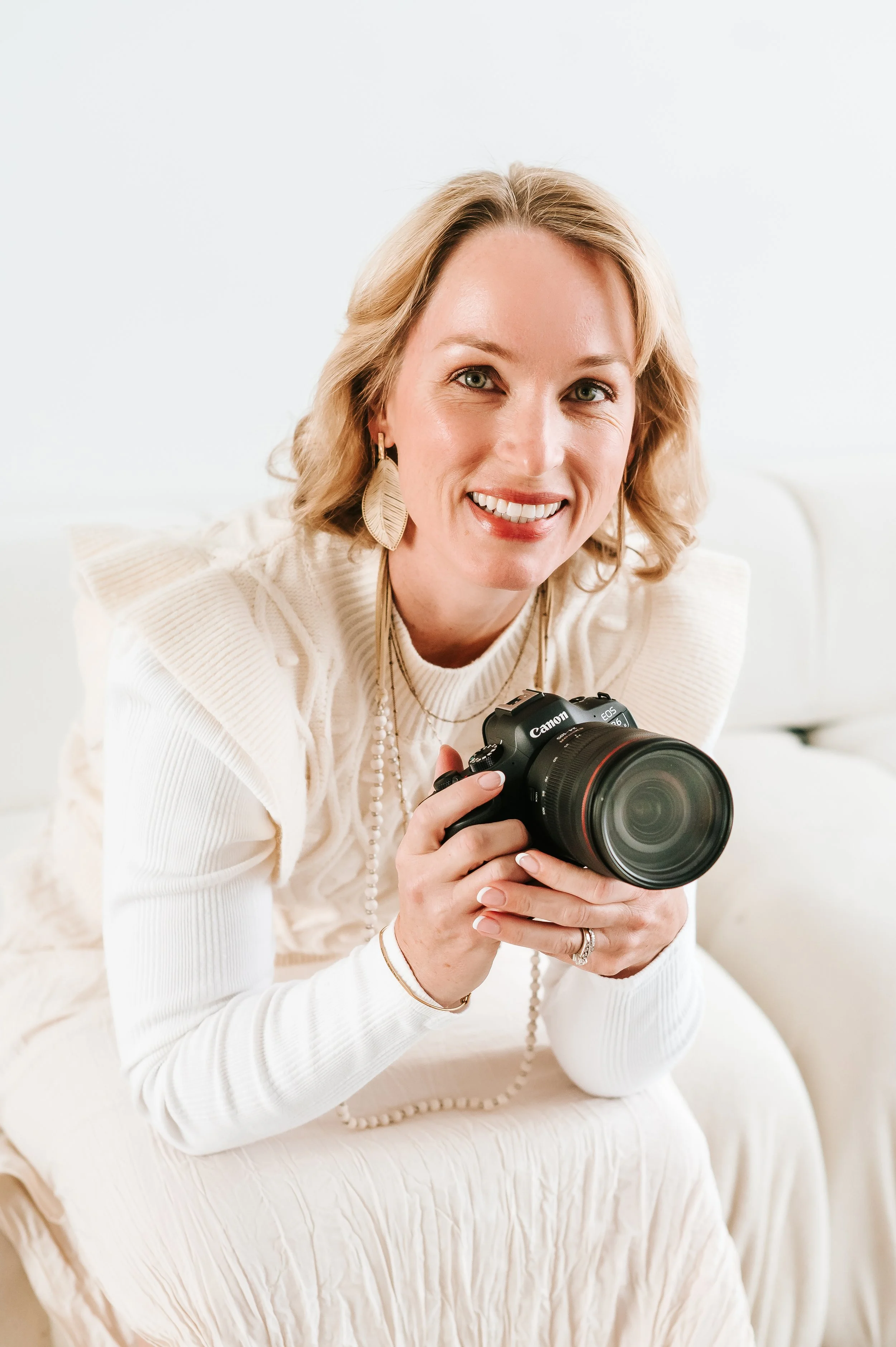 A woman with blonde hair smiling and holding a Canon camera, sitting on a light-colored couch in a bright, white room.