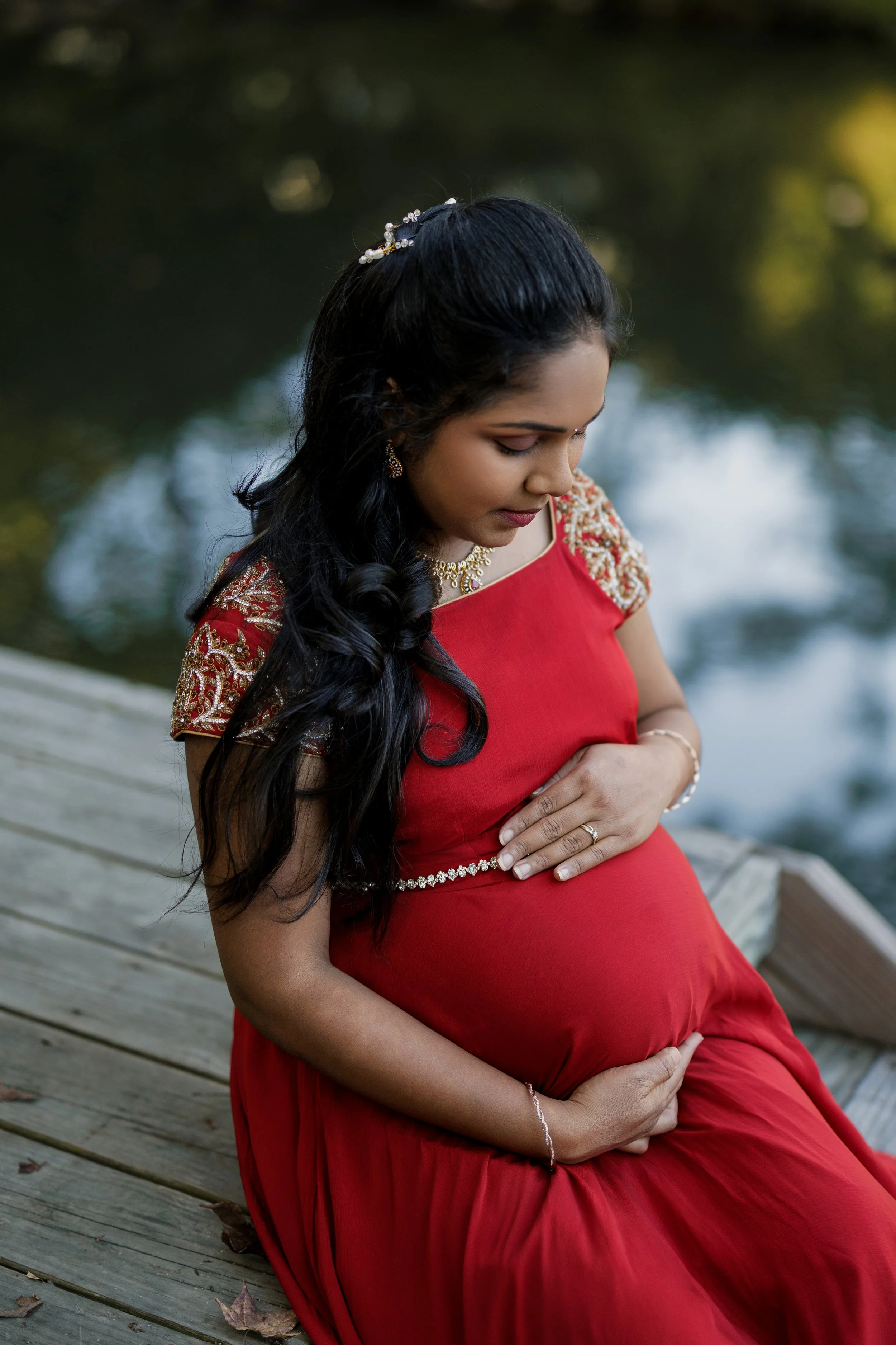 Pregnant woman in a red dress sitting on a wooden dock by a river, looking down at her belly with her hands gently resting on it.