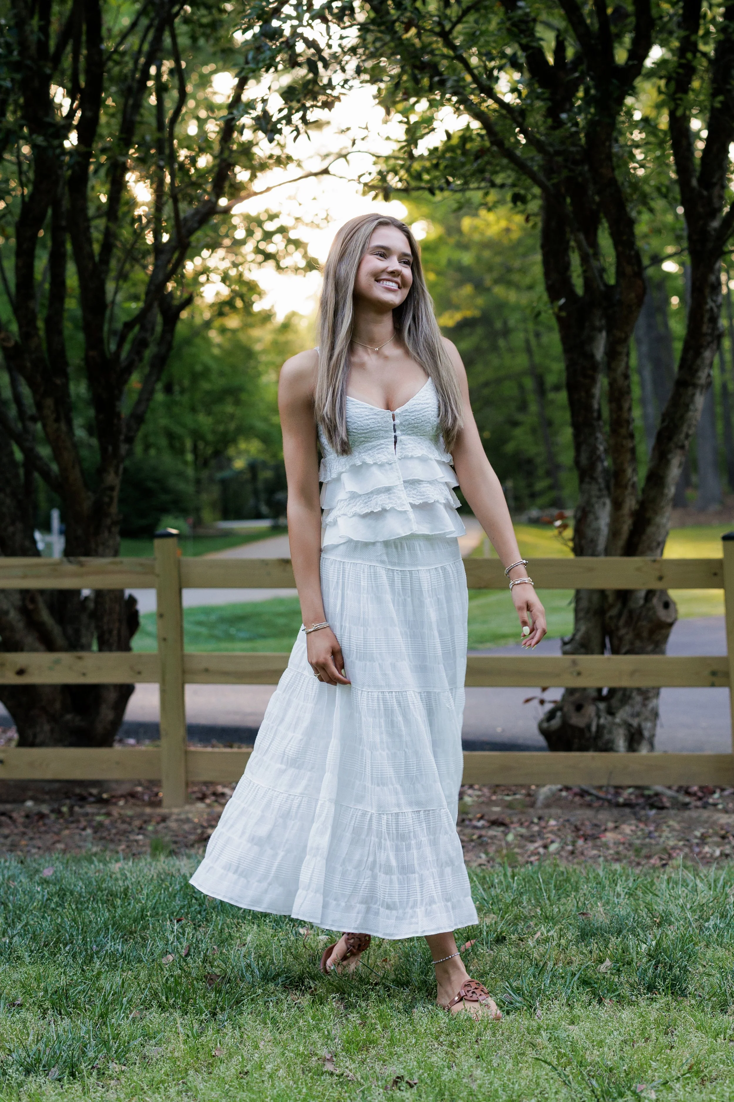 A young woman with long blonde hair in a white summer dress walking outdoors on grass with trees and a wooden fence in the background, smiling.