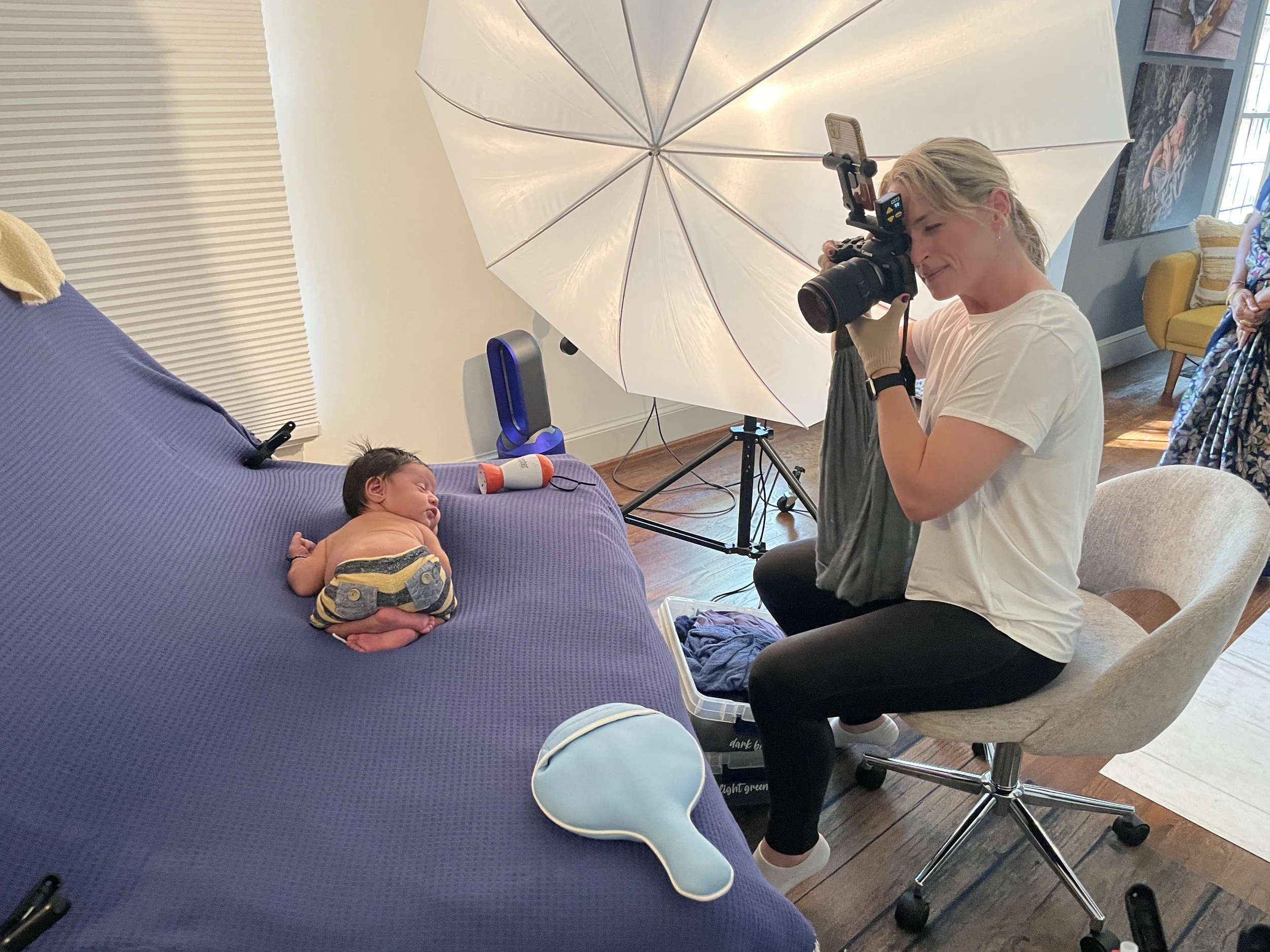 A DSLR photographer woman takes a portrait of a sleeping baby on a purple blanket, using a large umbrella for lighting in a cozy room with wooden floors and artwork on the walls.