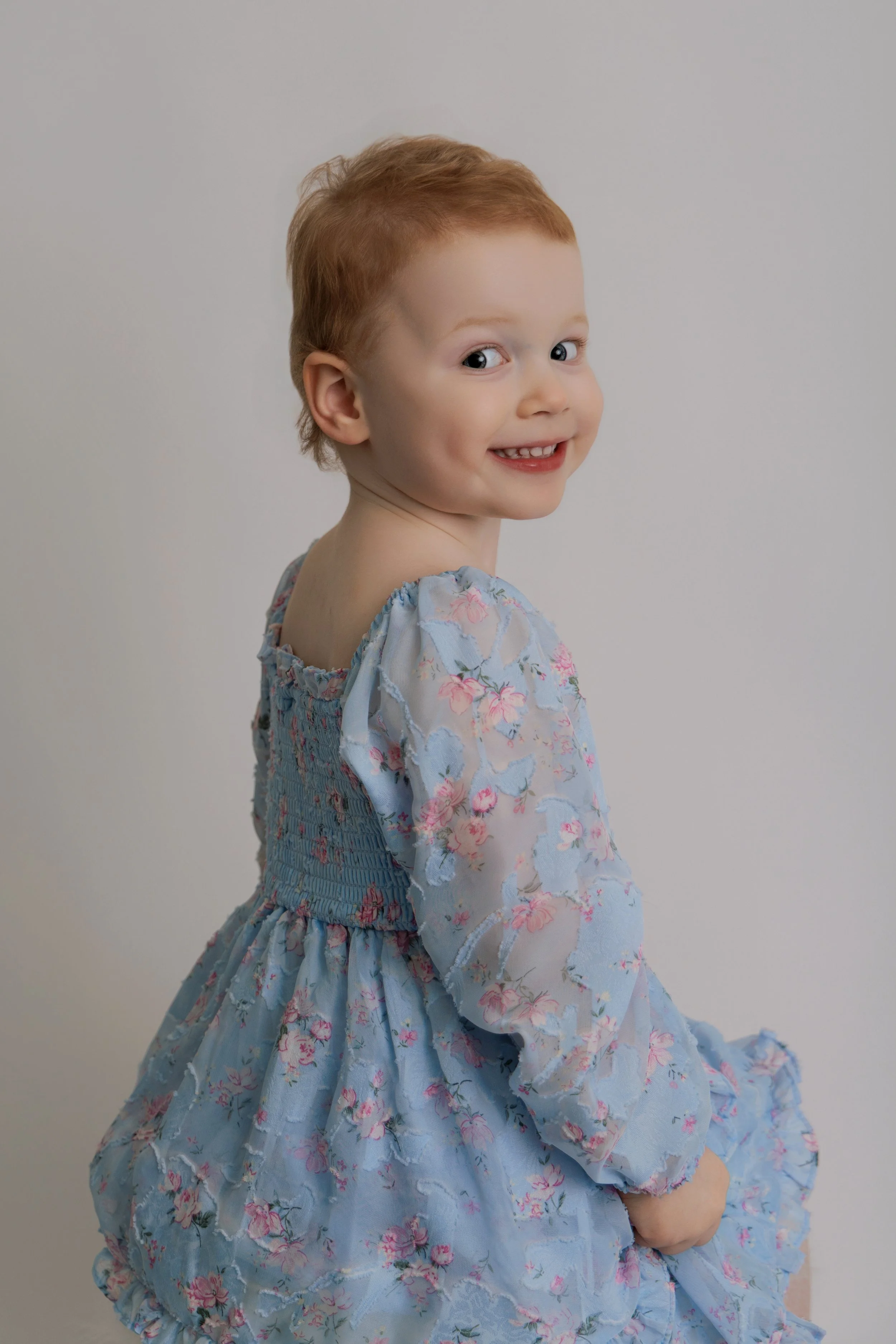 A young child with red hair sitting and looking over their shoulder, wearing a light blue floral dress, smiling at the camera.