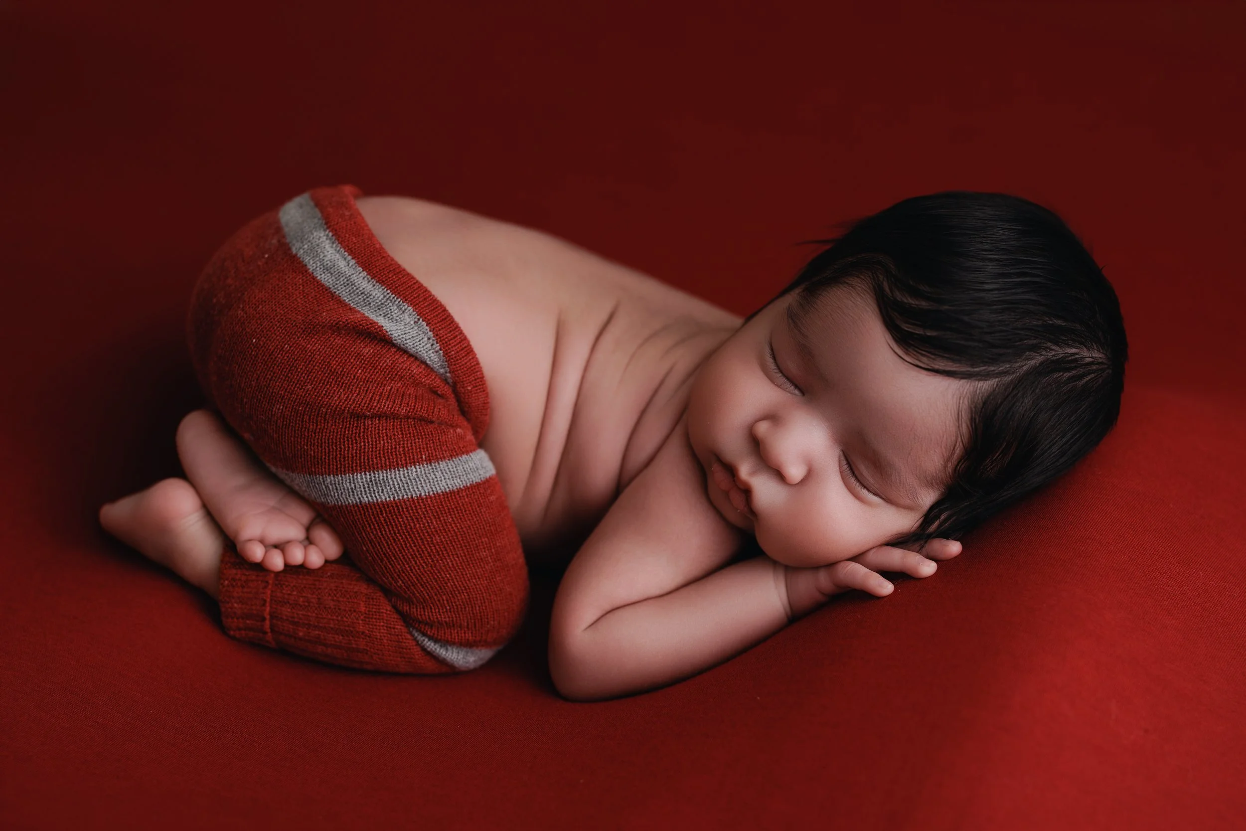 A baby with dark hair sleeping on a red background, wearing a red and gray striped shirt.