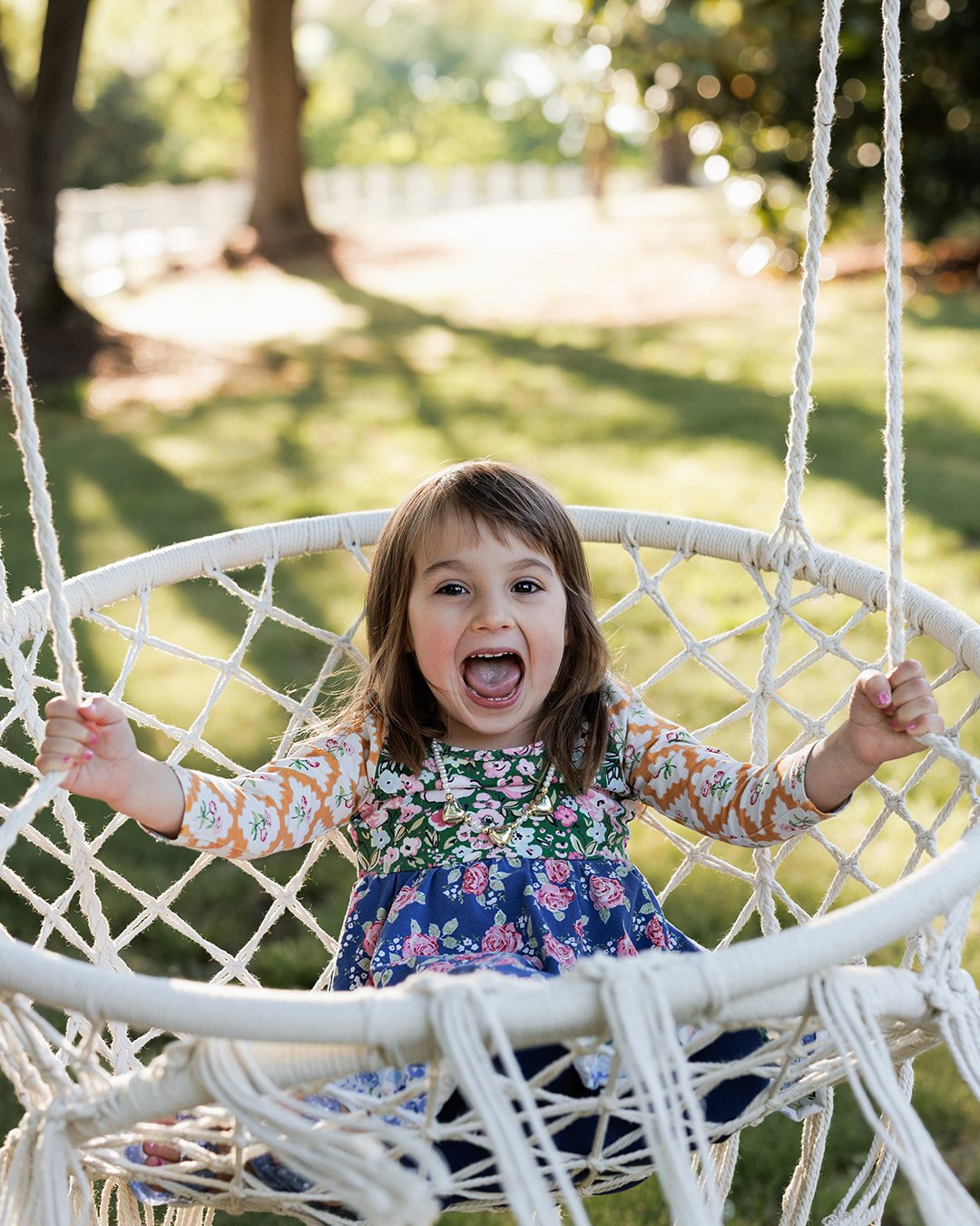 A young girl with long brown hair and a colorful floral dress laughing and sitting in a white woven hammock outdoors in a park or garden.