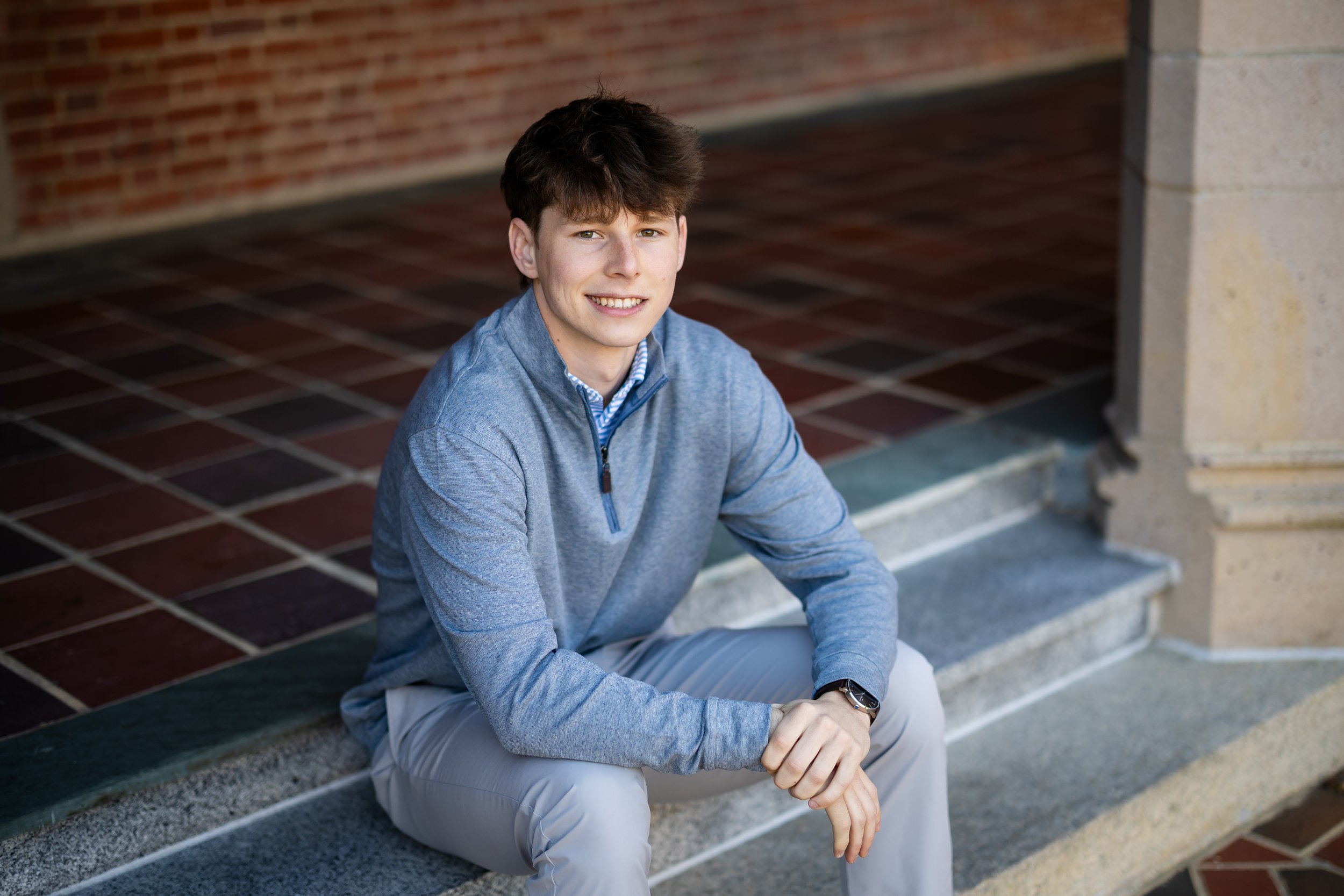 A young man with brown hair and a light smile, sitting on steps outside a brick building, wearing a light blue zip-up jacket and beige pants.