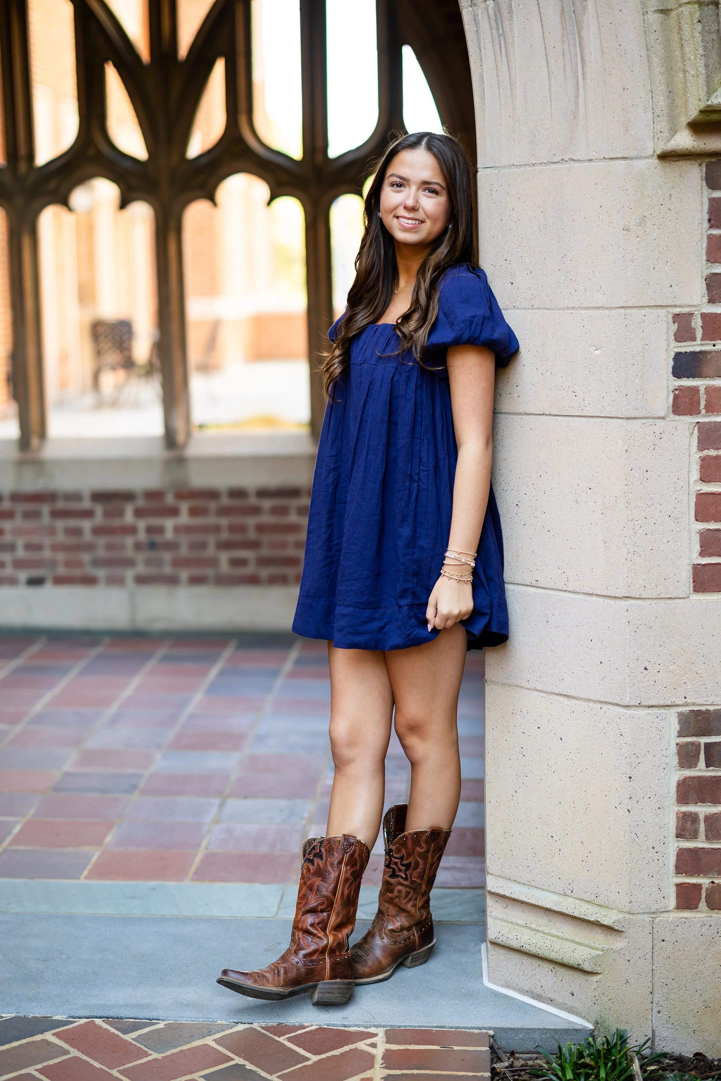 Young woman with long dark hair leaning against a brick building exterior with decorative architectural elements, wearing a blue dress and cowboy boots.