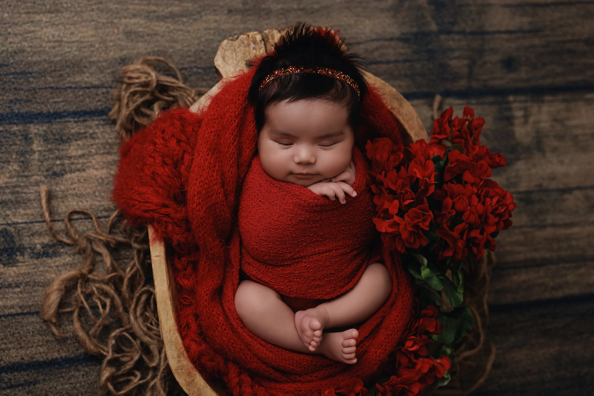 A sleeping baby wrapped in red cloth with a red headband, lying in a wooden basket surrounded by red flowers on a rustic wooden surface.