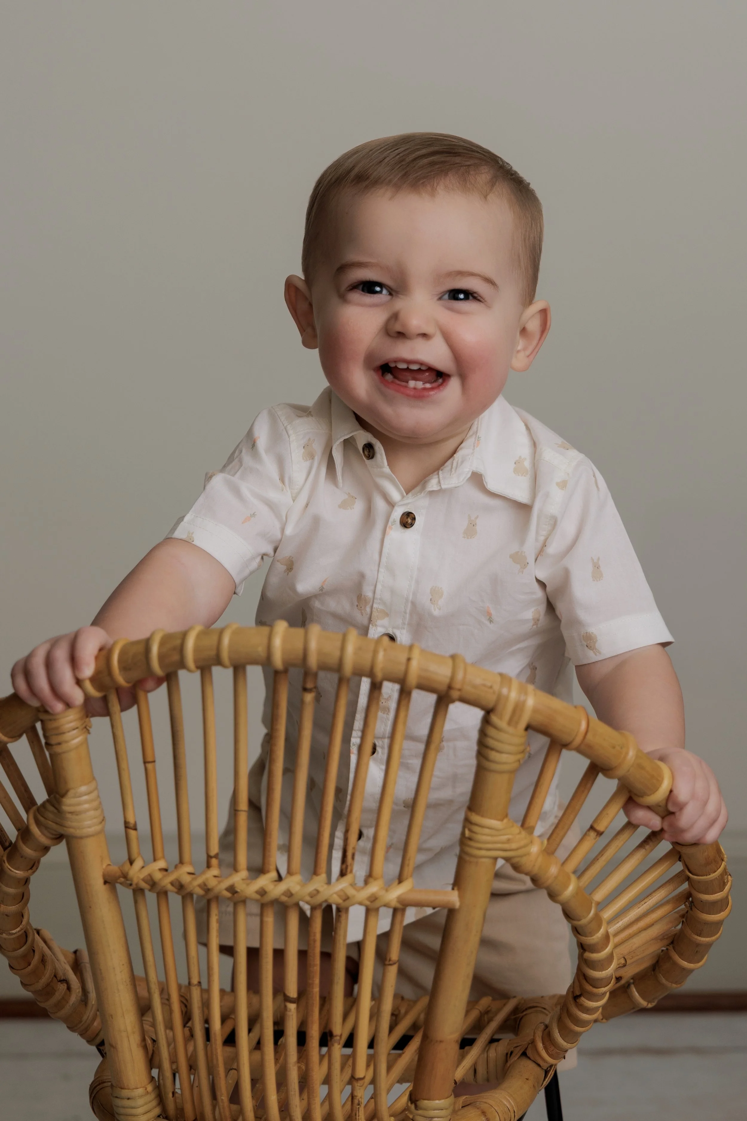 Smiling young boy with short hair and blue eyes, wearing a light-colored shirt with bunny prints, holding onto a rattan chair in front of a plain background.