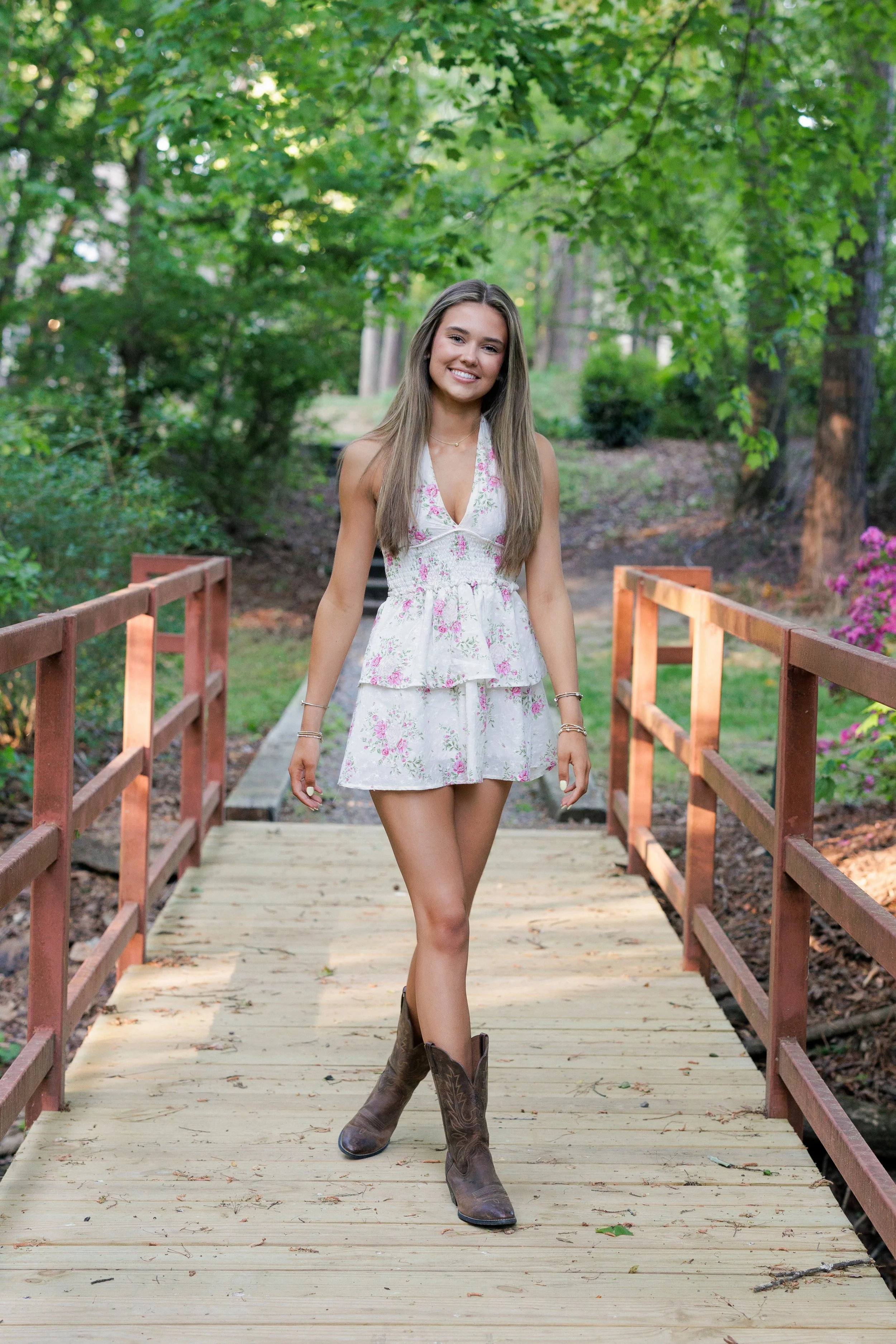 A young woman with long brown hair wearing a white floral dress and cowboy boots walking on a wooden bridge in a lush green park.