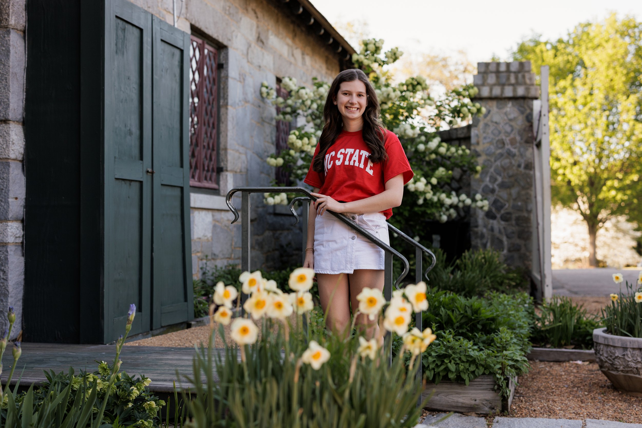 A young woman with long dark hair, smiling, wearing a red Ohio State t-shirt and a white skirt, standing next to a metal handrail in a garden with yellow and white flowers, a stone building with green shutters, and trees in the background during dayt