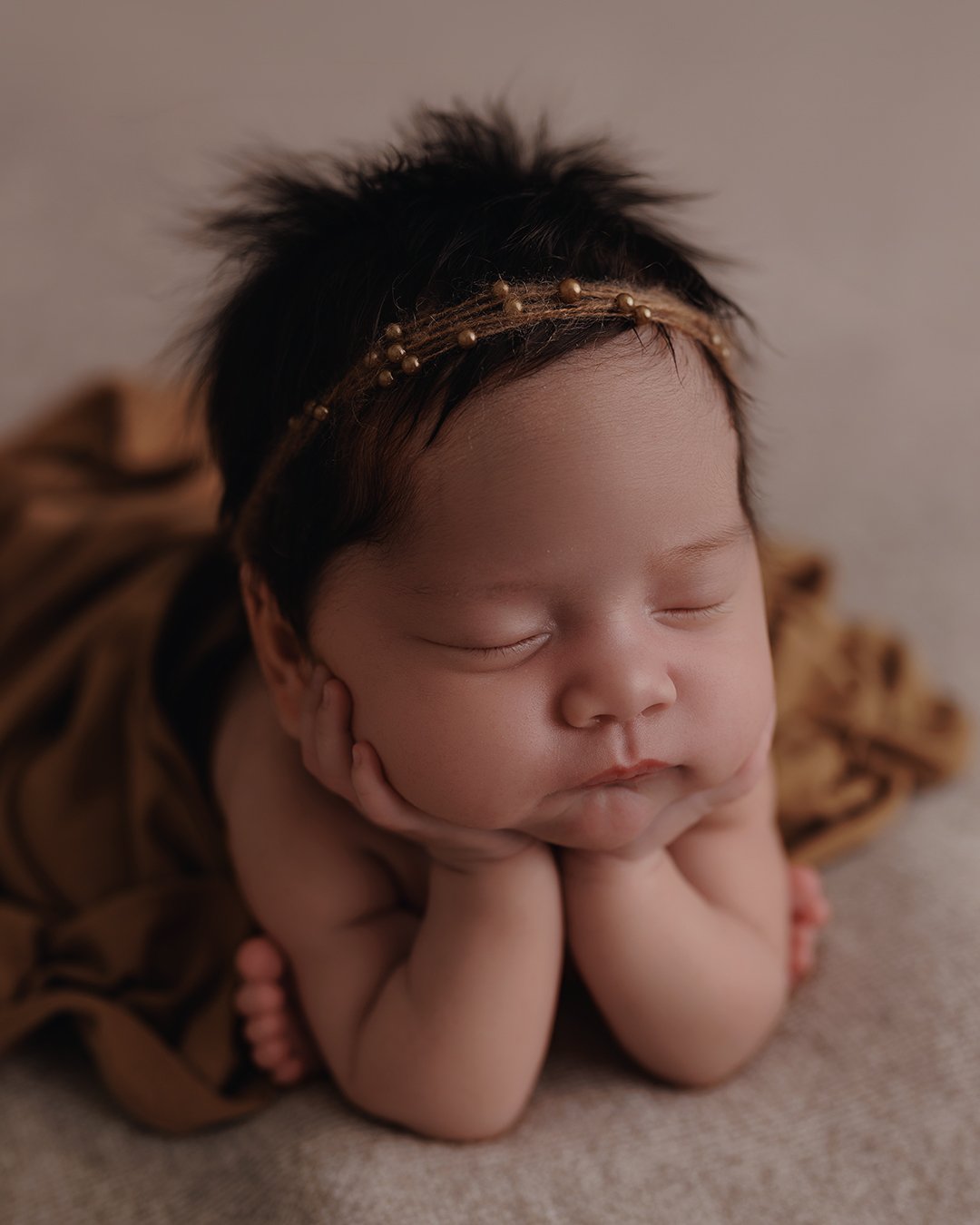 Close-up of a sleeping newborn baby with hands resting on cheeks, wearing a gold headband and a brown patterned outfit.